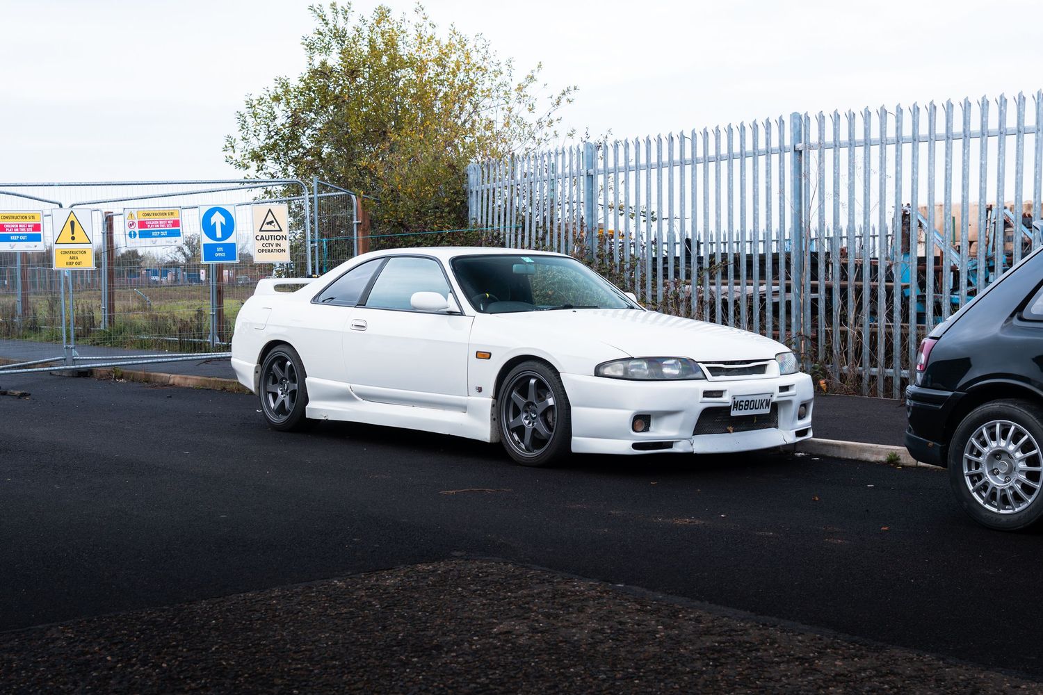 White Nissan Skyline R33 GT-R parked in a lot with road signs and metal fencing behind it.
