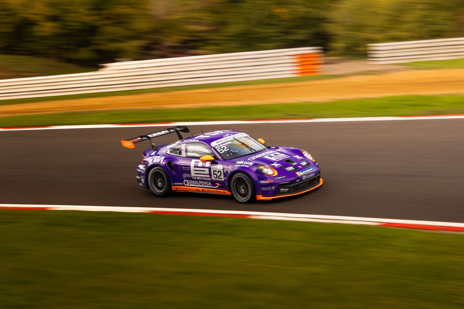 Purple Porsche 911 GT3 Cup race car cornering on a track with warm autumn trees in the background.