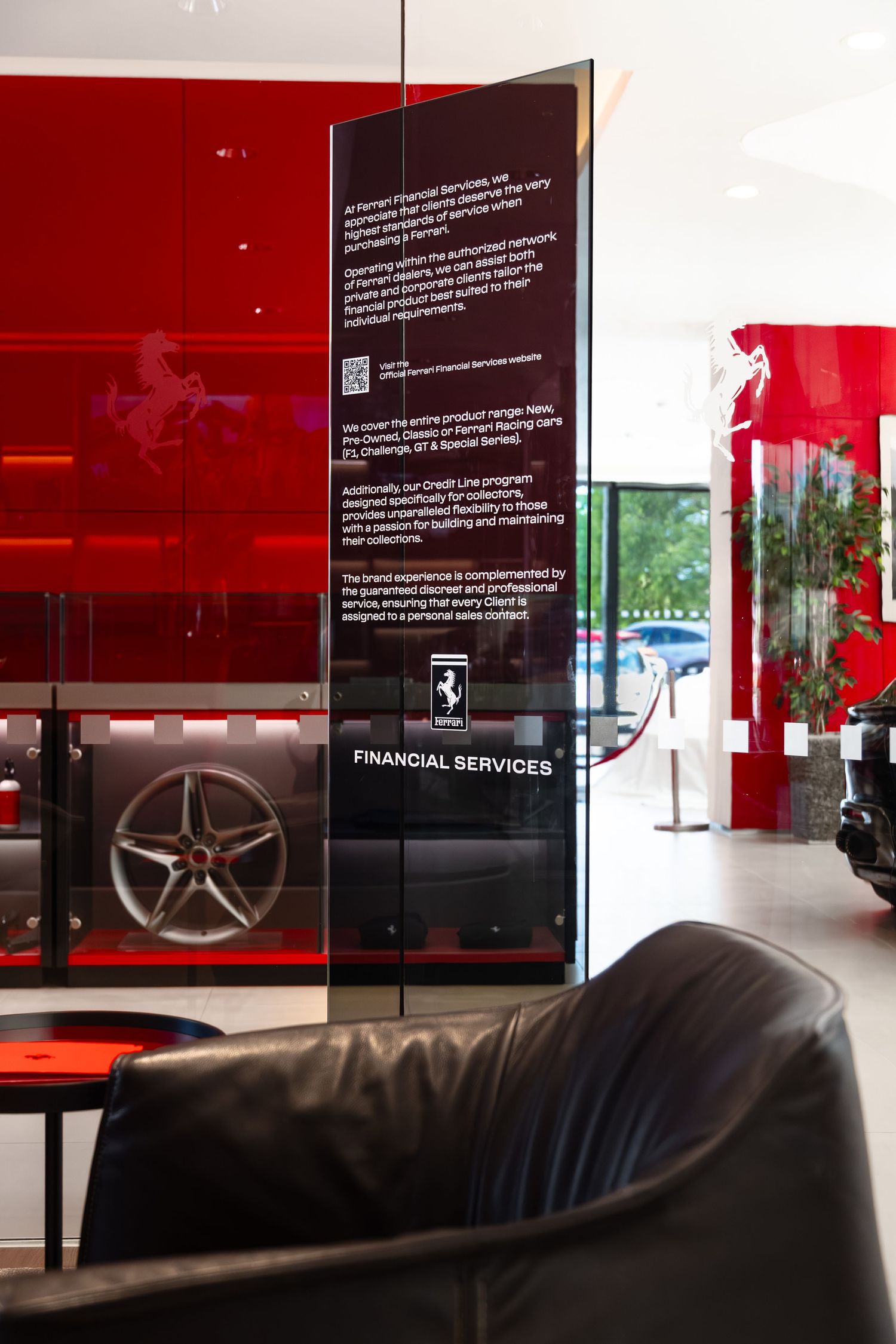 Ferrari dealership showroom featuring Financial Services signage, red walls, luxury seating, and a display of a Ferrari alloy wheel.