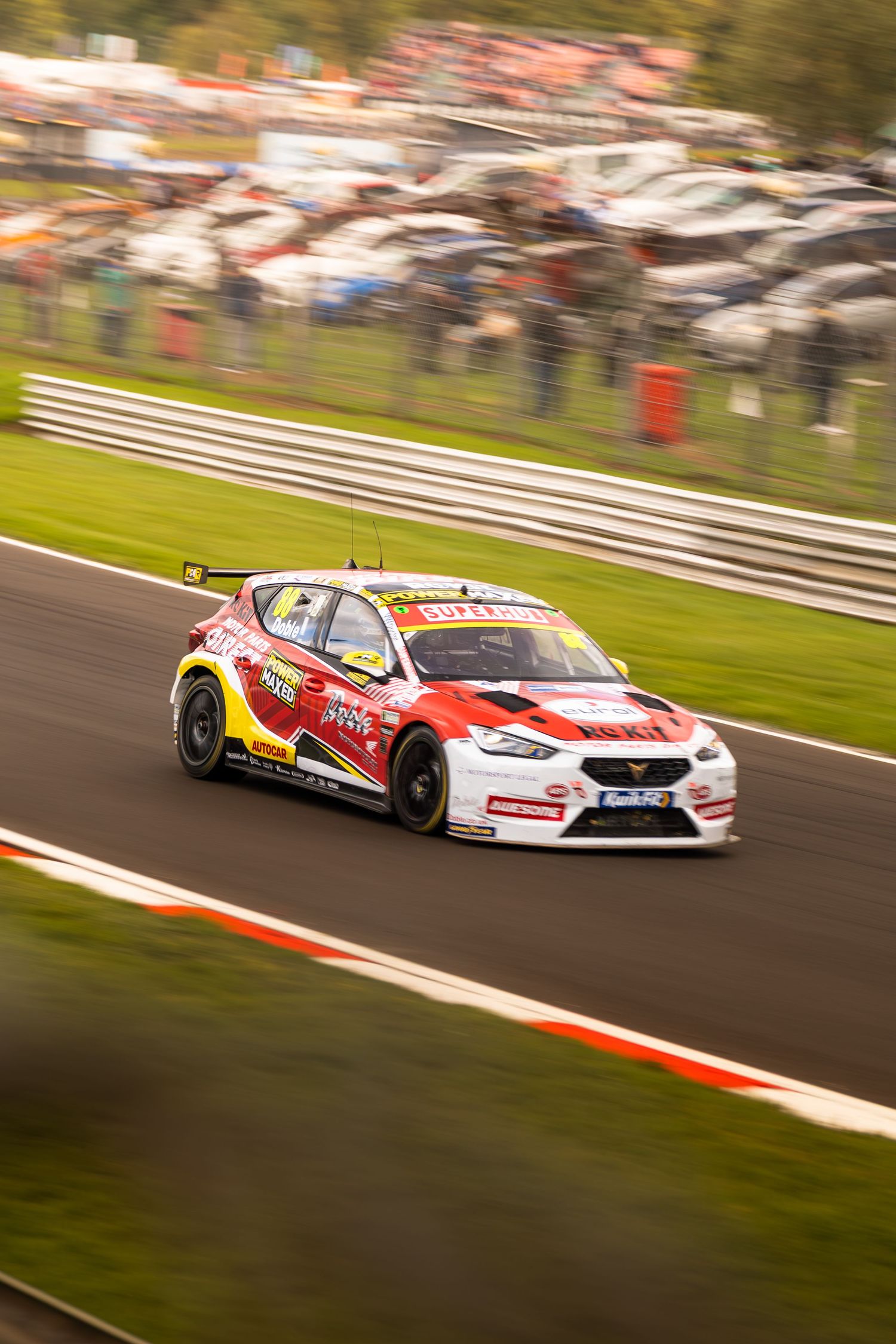A red and yellow CUPRA touring car races at speed through a corner at a British motorsport circuit.