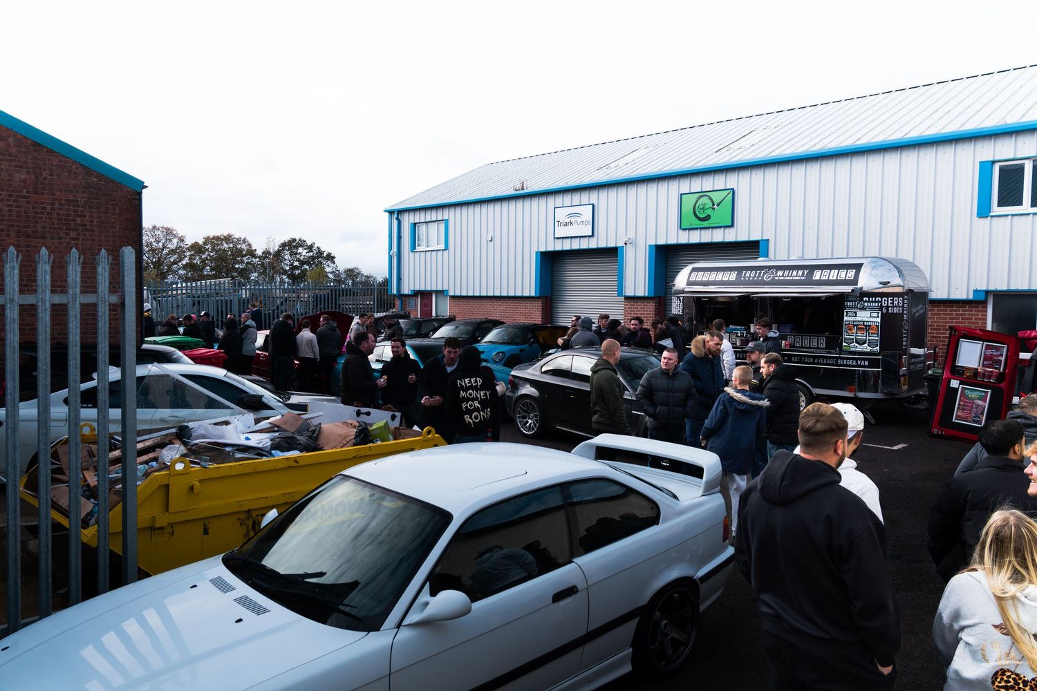 Large crowd gathered outside an automotive workshop unit at an outdoor car meet event.