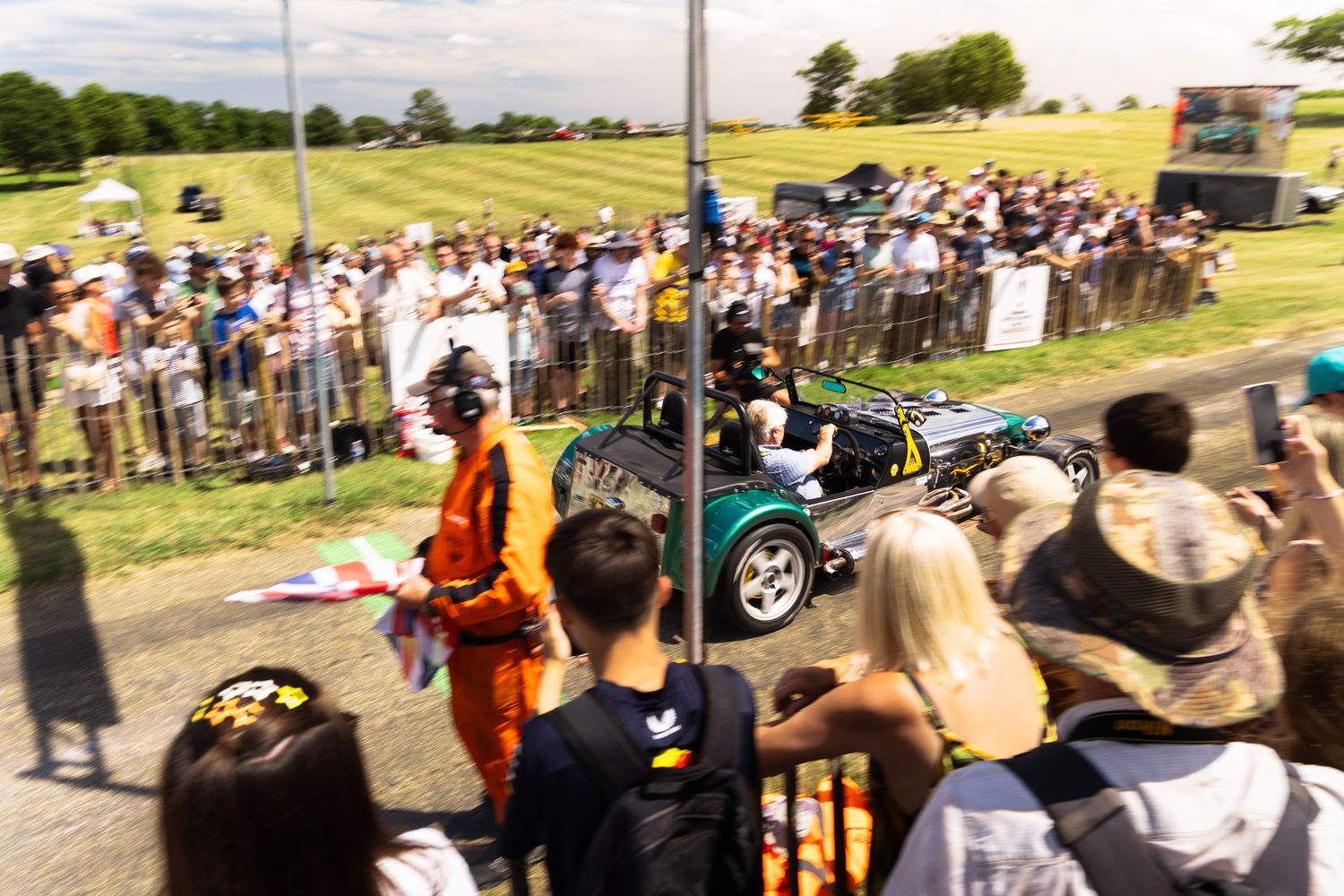 Green open-top race car drives past cheering crowd at outdoor motorsport event on a sunny day.
