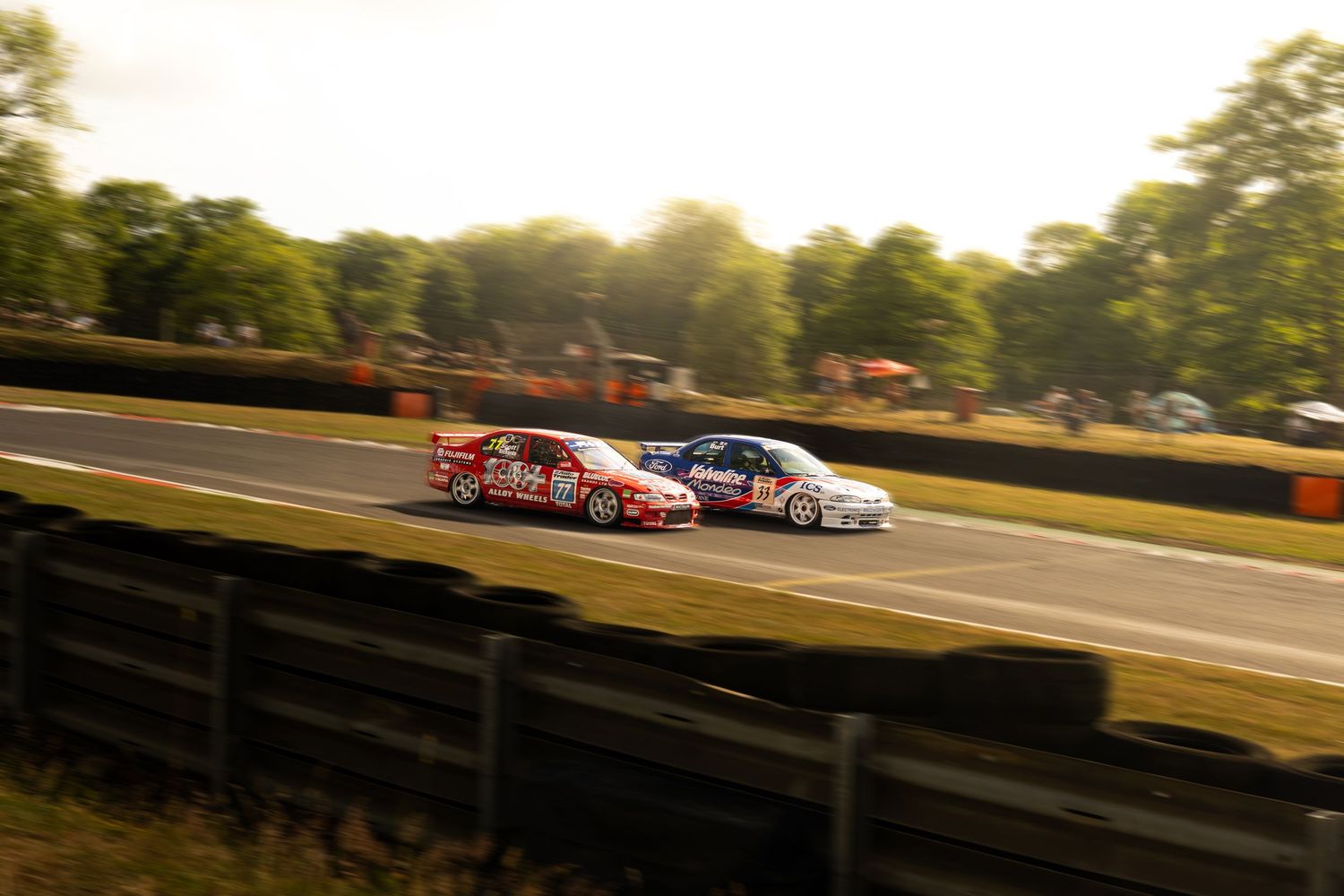 Two BTCC touring cars racing side by side on tree-lined circuit in golden evening light.