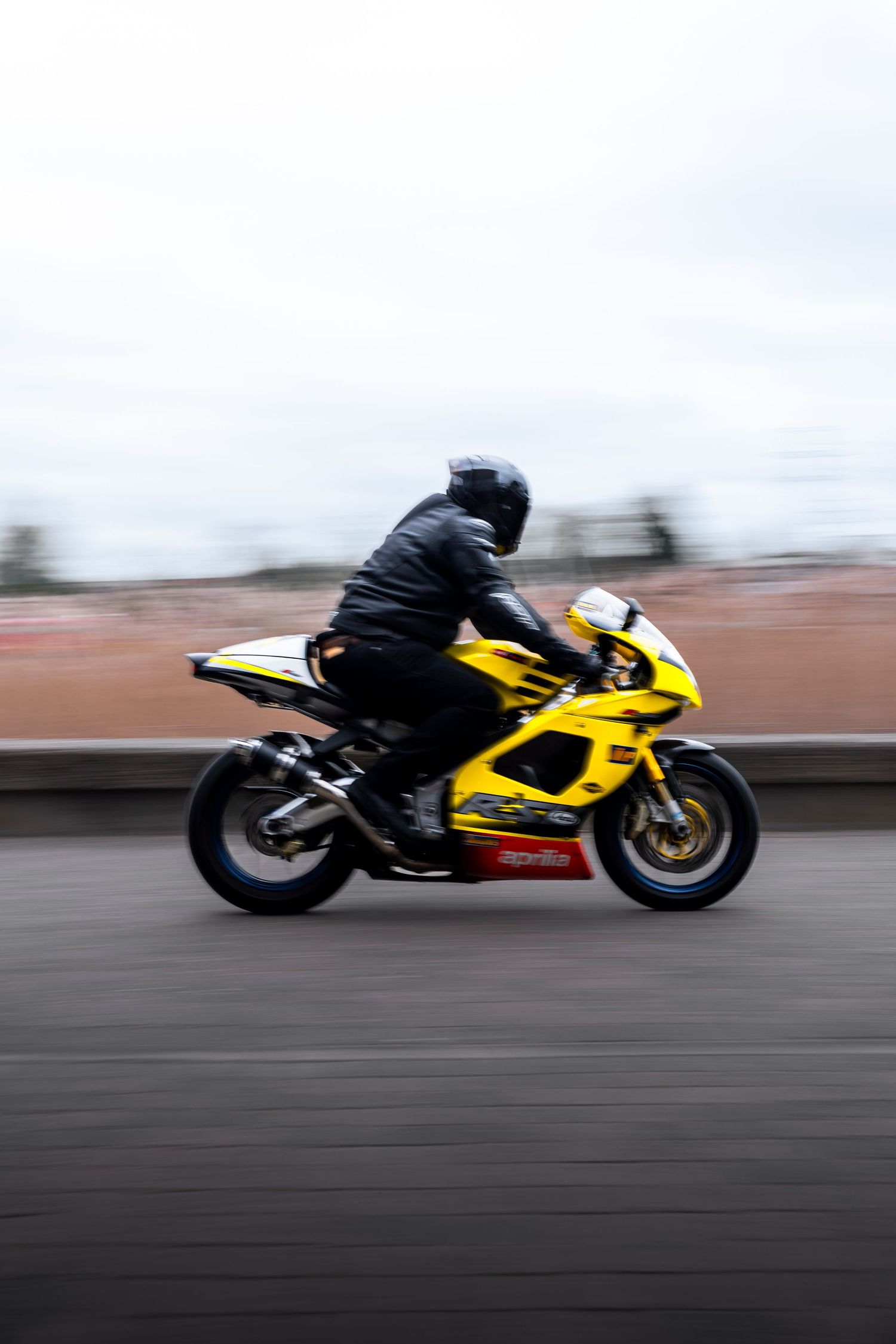 Panning shot of a yellow Aprilia RSV superbike with motion blur, rider in black gear.
