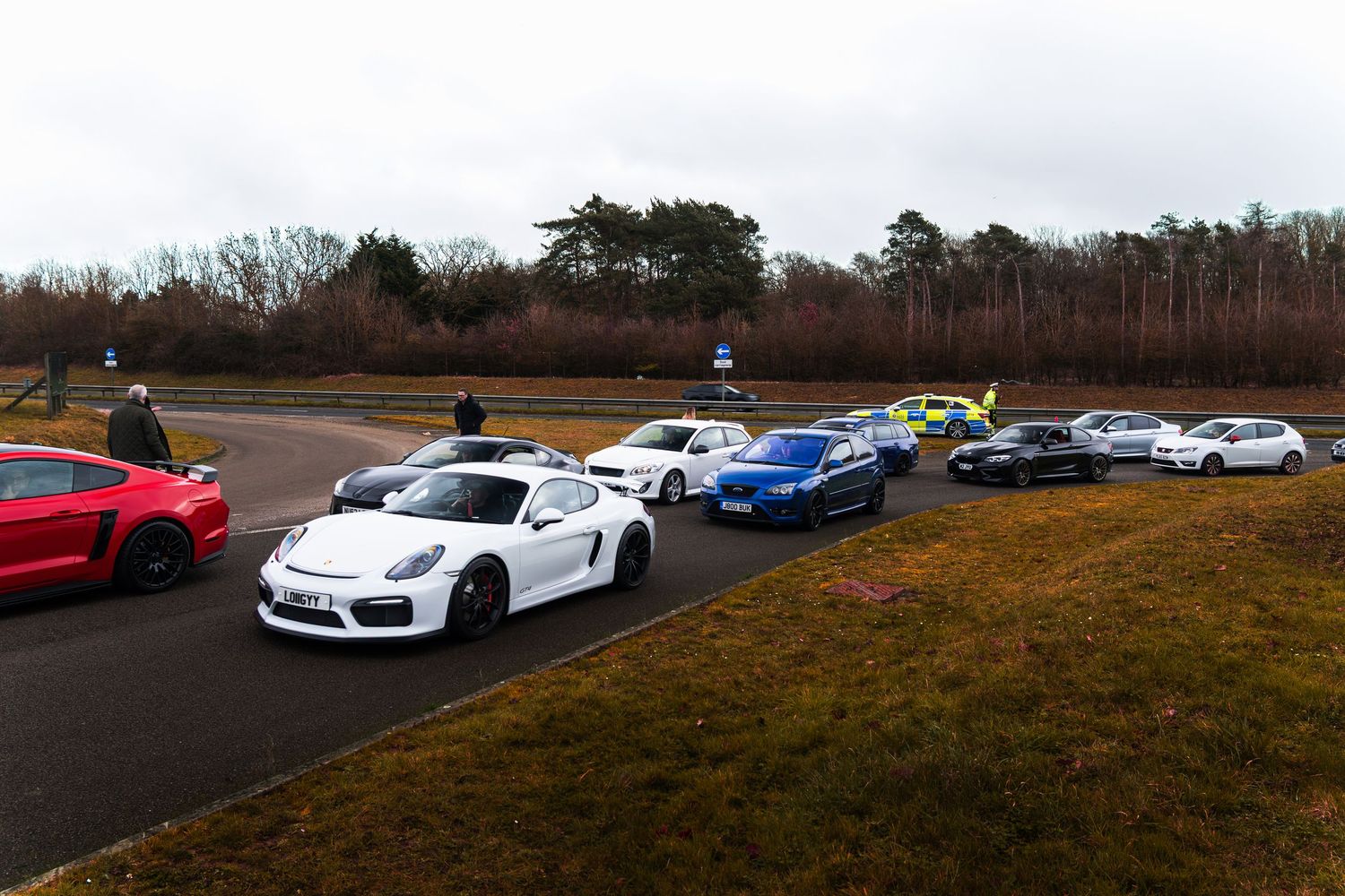 A white Porsche Cayman GT4 leads a lineup of sports cars on a track at a car meet.