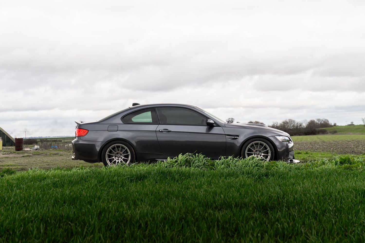 Grey BMW M3 coupe parked beside green field with cloudy sky in rural landscape.