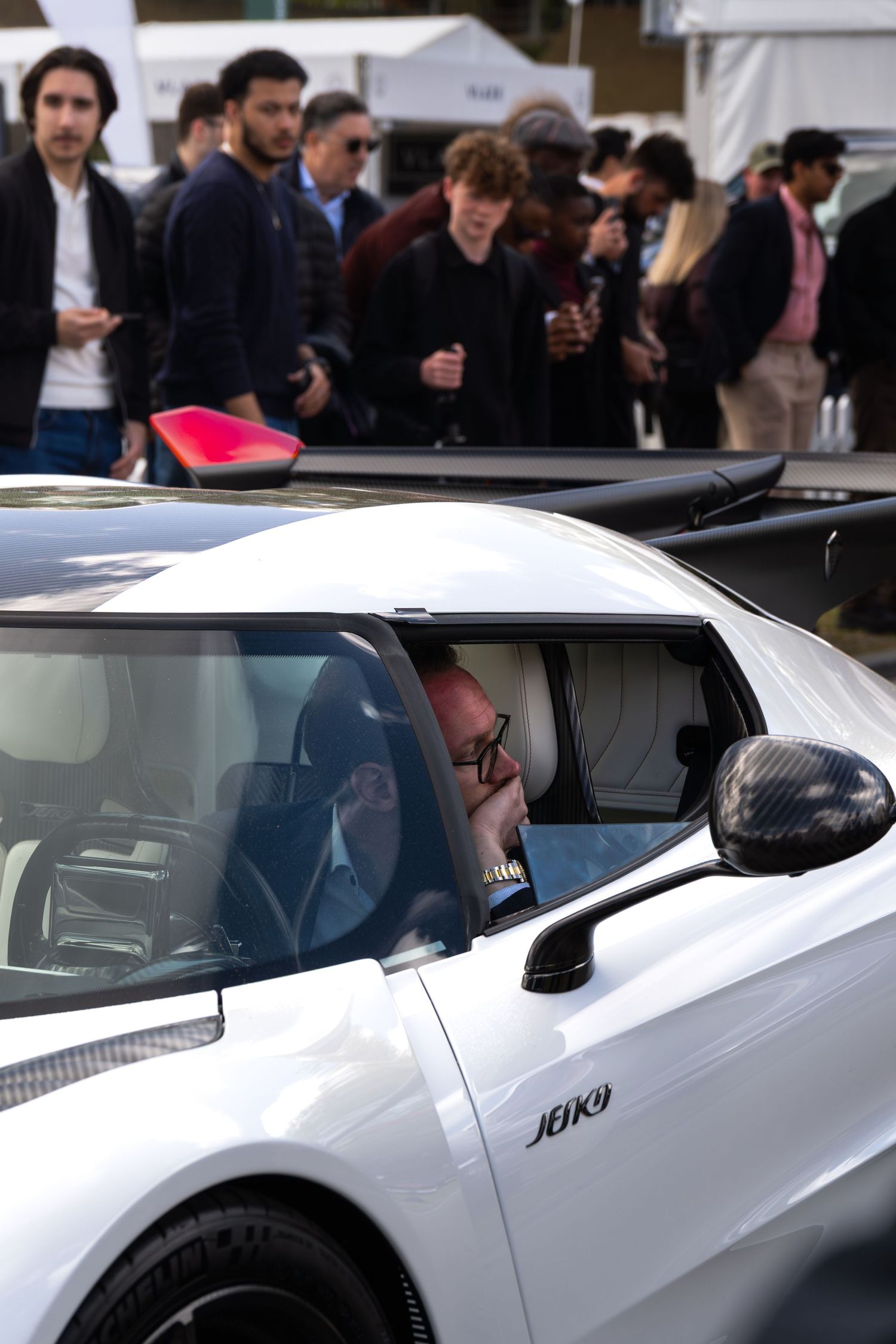 Man seated inside a white supercar at an outdoor car show event with crowd in background.
