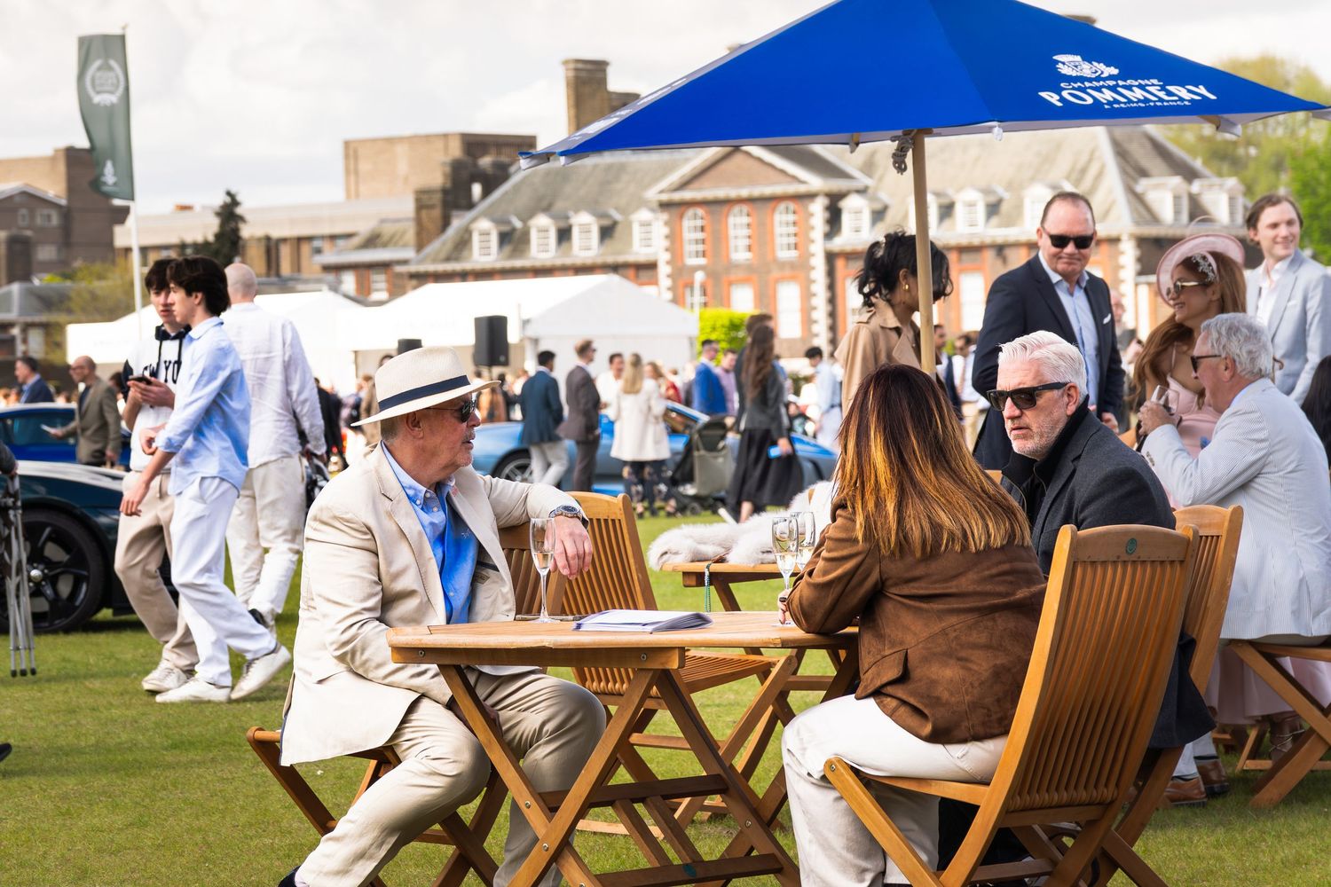 Elegantly dressed guests relax at outdoor wooden tables under a Koenigsegg umbrella at a luxury car event.