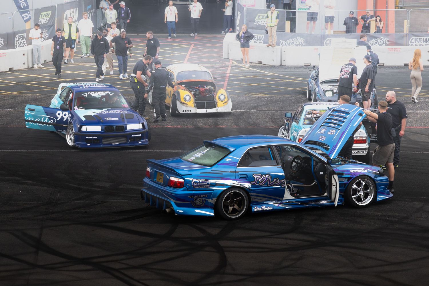 Multiple blue drift cars including a Nissan Skyline are parked on a black asphalt track at a motorsport event.