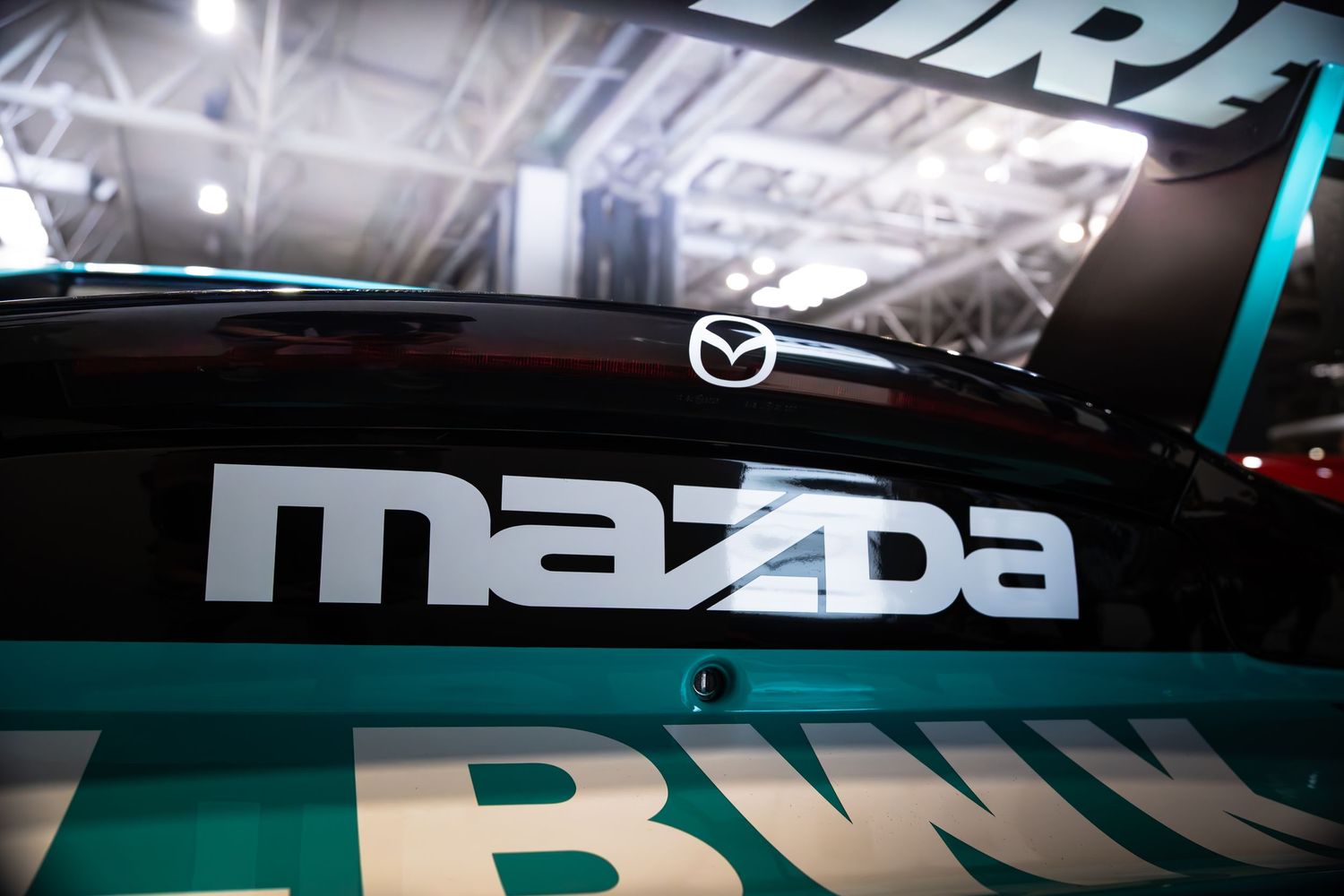 Mazda logo and branding on the rear window of a teal racing car at an auto show.
