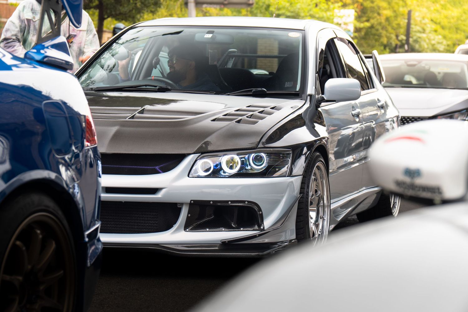 Modified silver Mitsubishi Lancer Evolution with angel eyes surrounded by cars at a meet.