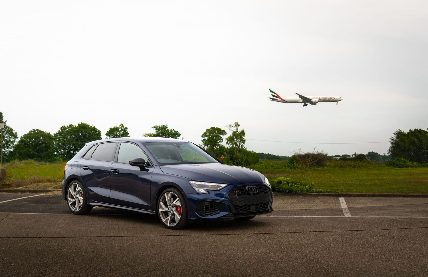 Navy blue Audi S3 Sportback parked outdoors with a commercial airplane flying overhead against cloudy sky.