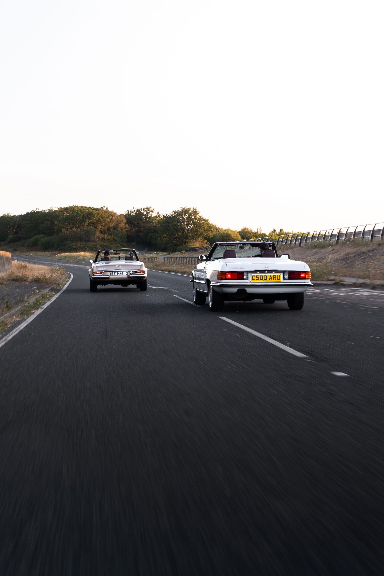 Two white classic convertible cars driving on a winding road surrounded by trees and hills at dusk.