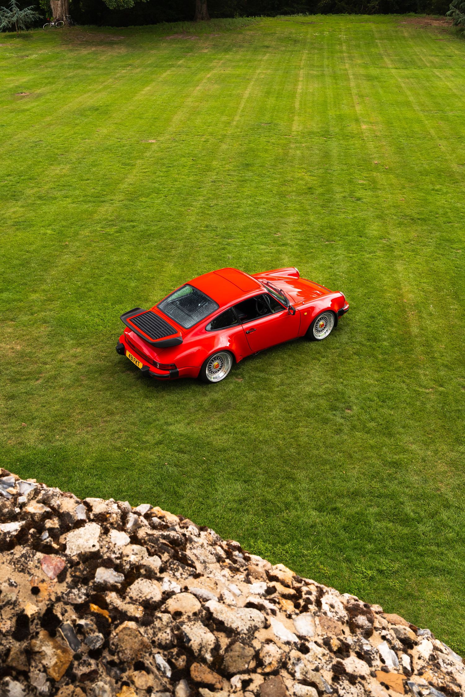 Red Porsche 911 Turbo parked on lush striped green lawn, aerial view with stone wall foreground.
