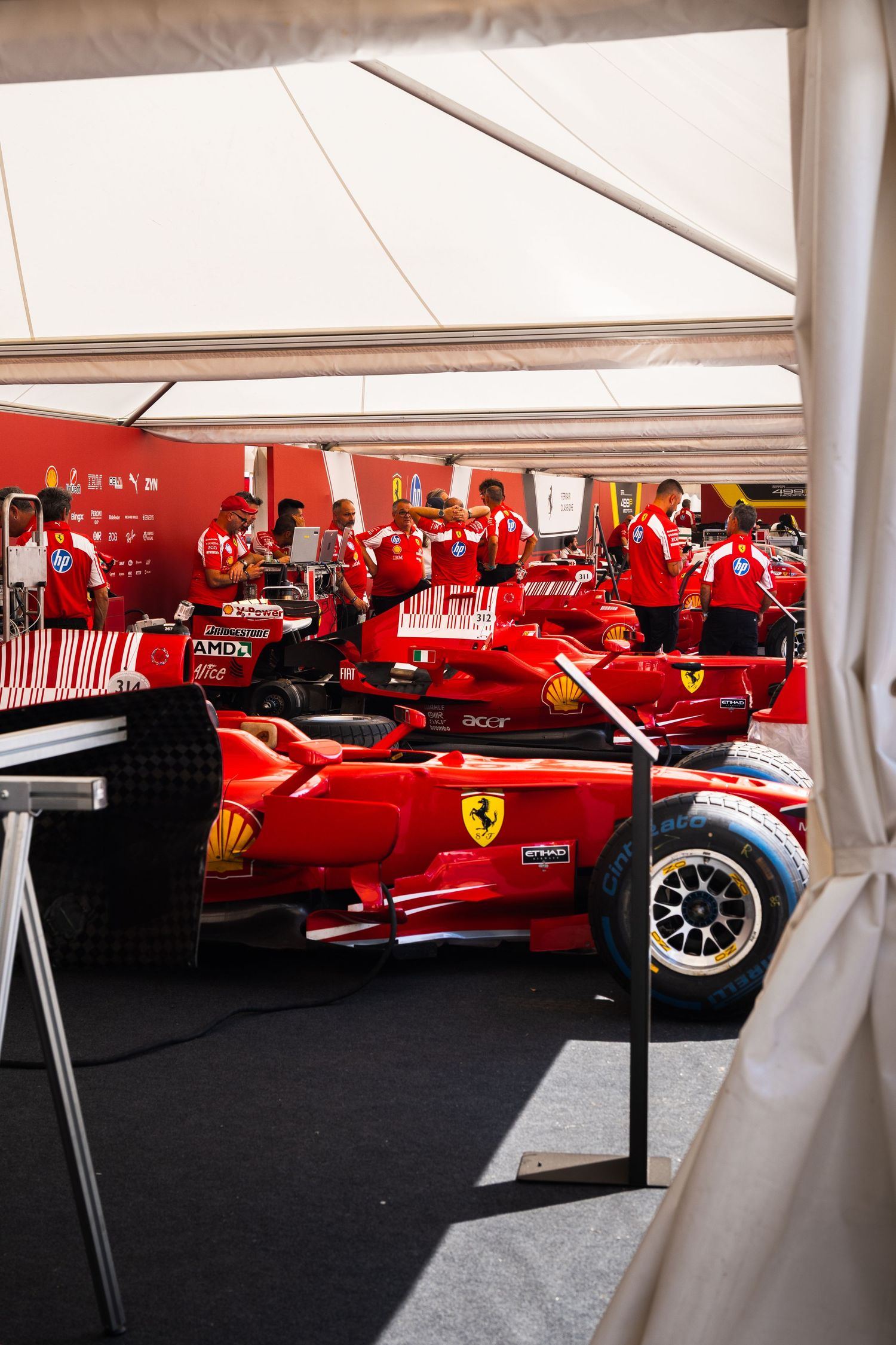 Ferrari F1 cars in red livery lined up in the paddock garage with mechanics in red uniforms working on them.