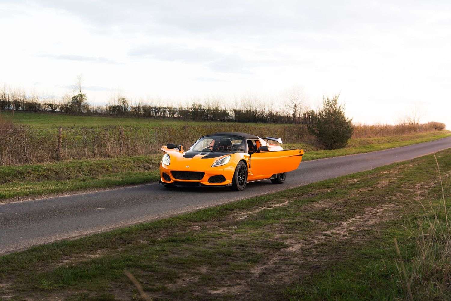 Orange Lotus Elise sports car driving on a rural road with green hedgerows at sunset.