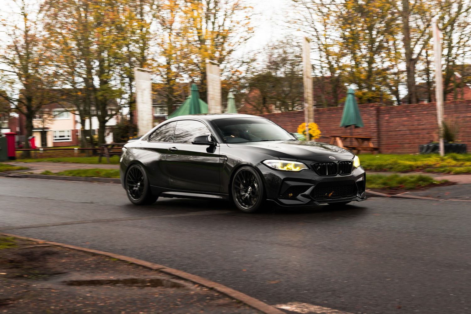 Black BMW M2 coupe driving through wet UK car park surrounded by autumn trees.