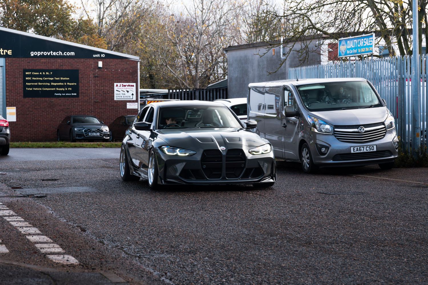 Black BMW M3 with aggressive wide body kit parked next to grey Vauxhall Vivaro van at garage.