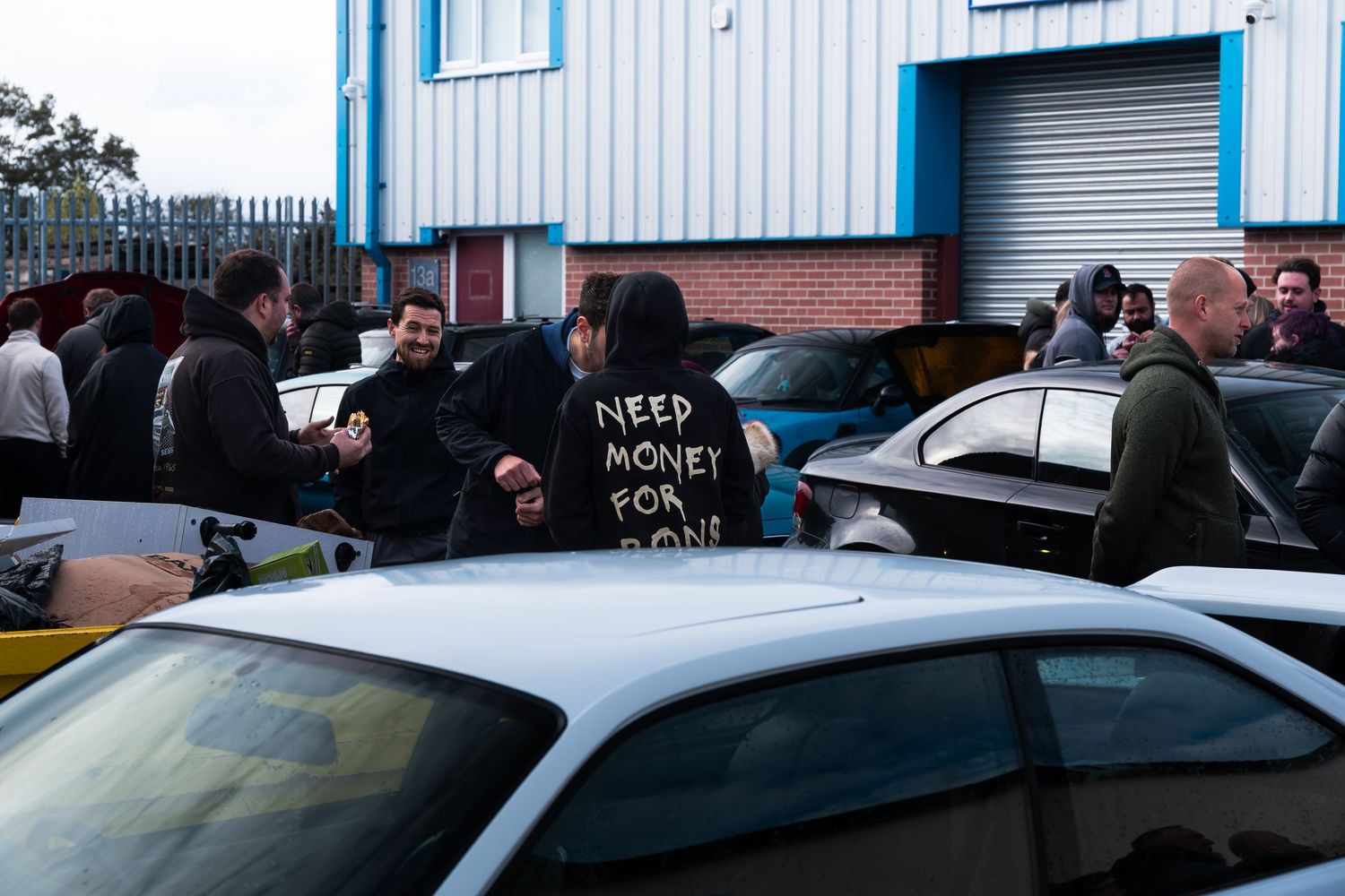 Group of people socializing at an outdoor car meet, one wearing a Need Money For Mods hoodie.