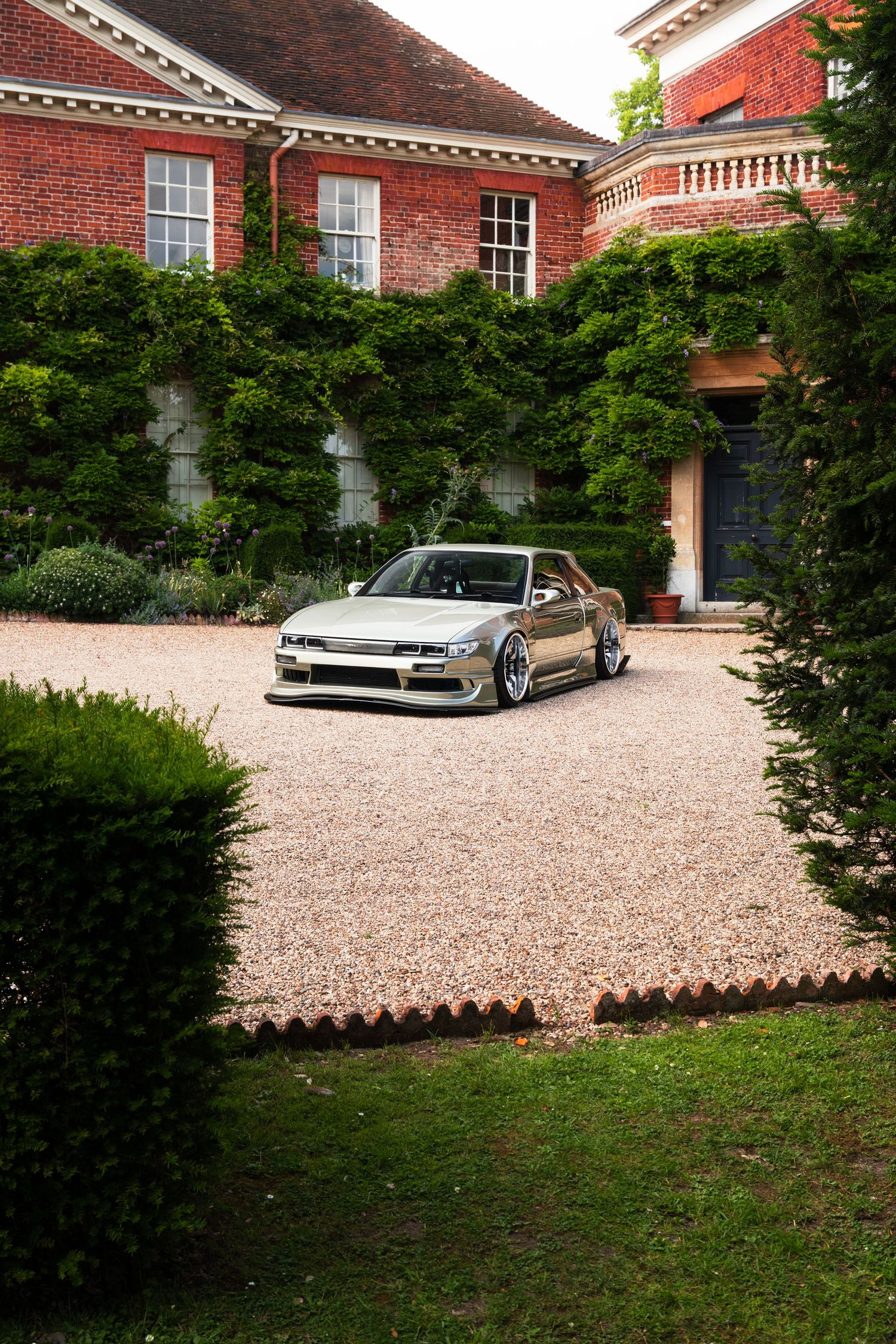 A silver modified Nissan Silvia S13 parked on a gravel driveway outside a red brick manor house.