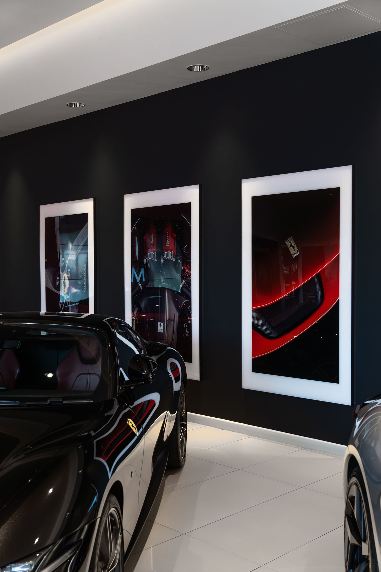 Luxury car showroom with black sports cars displayed against dark walls featuring framed automotive art prints.