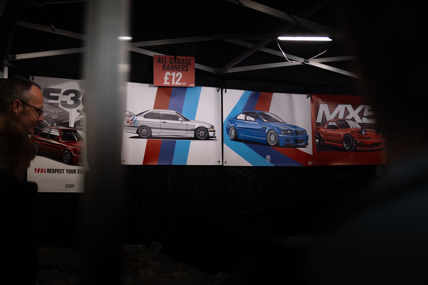 Car show vendor booth displaying E3, E46, and MX5 automotive posters priced at £12 each.