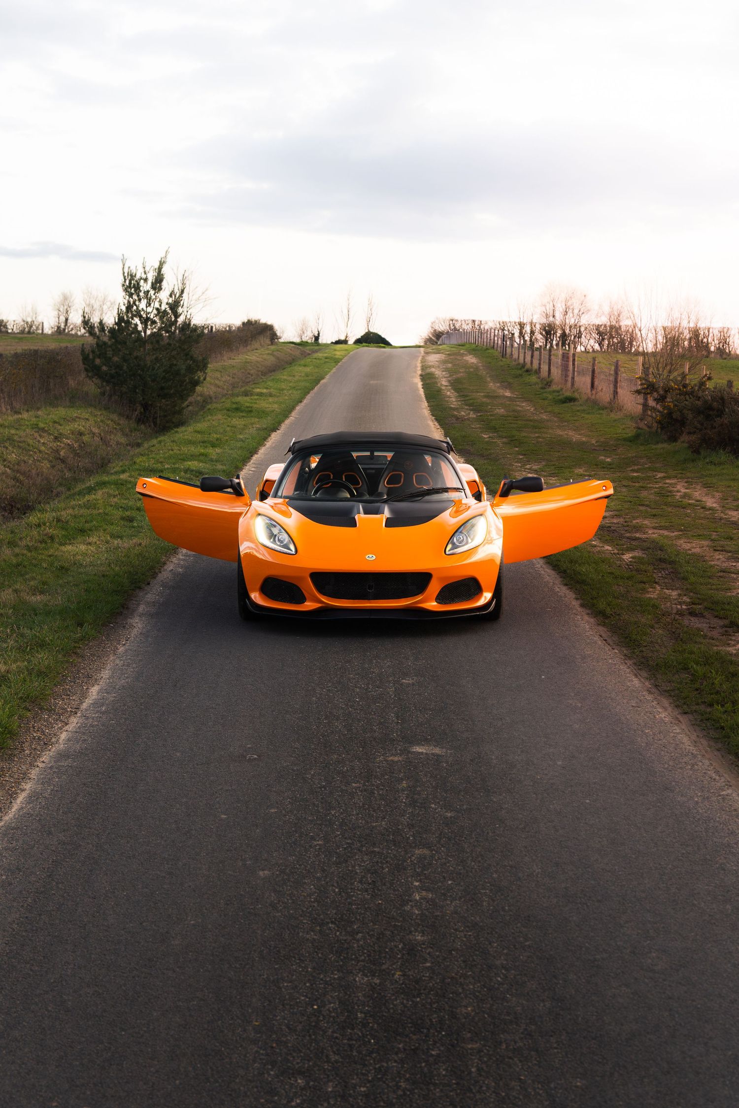 Orange Lotus Elise with doors open on a narrow road flanked by trees and green fields.
