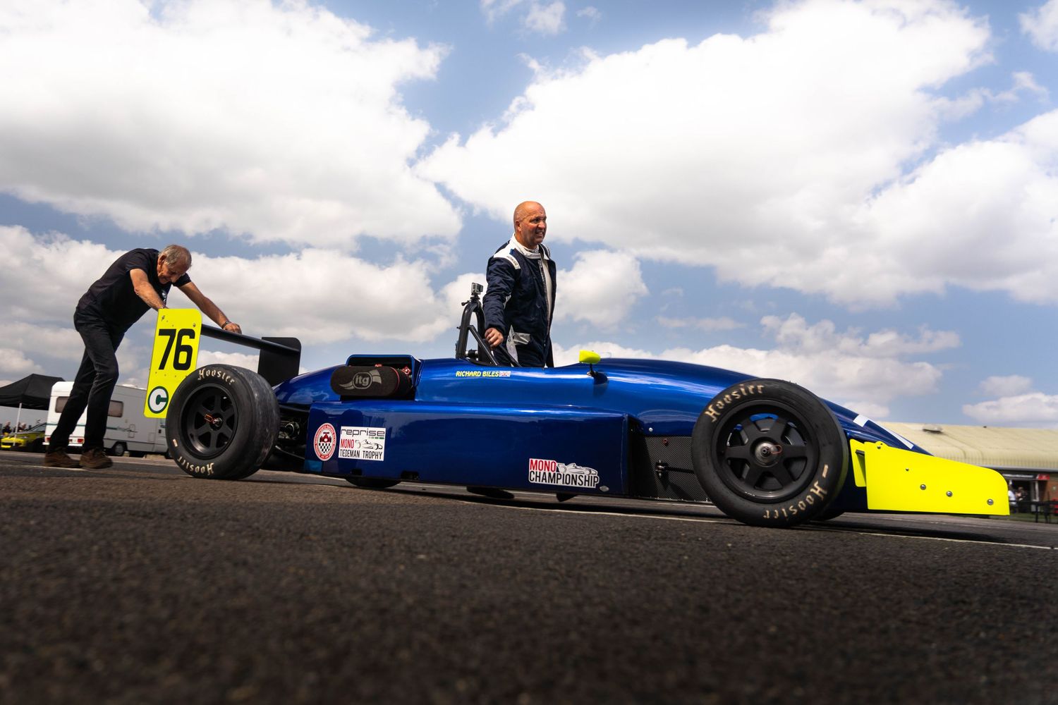 A driver stands beside blue Formula 76 racing car on the track under a cloudy sky.