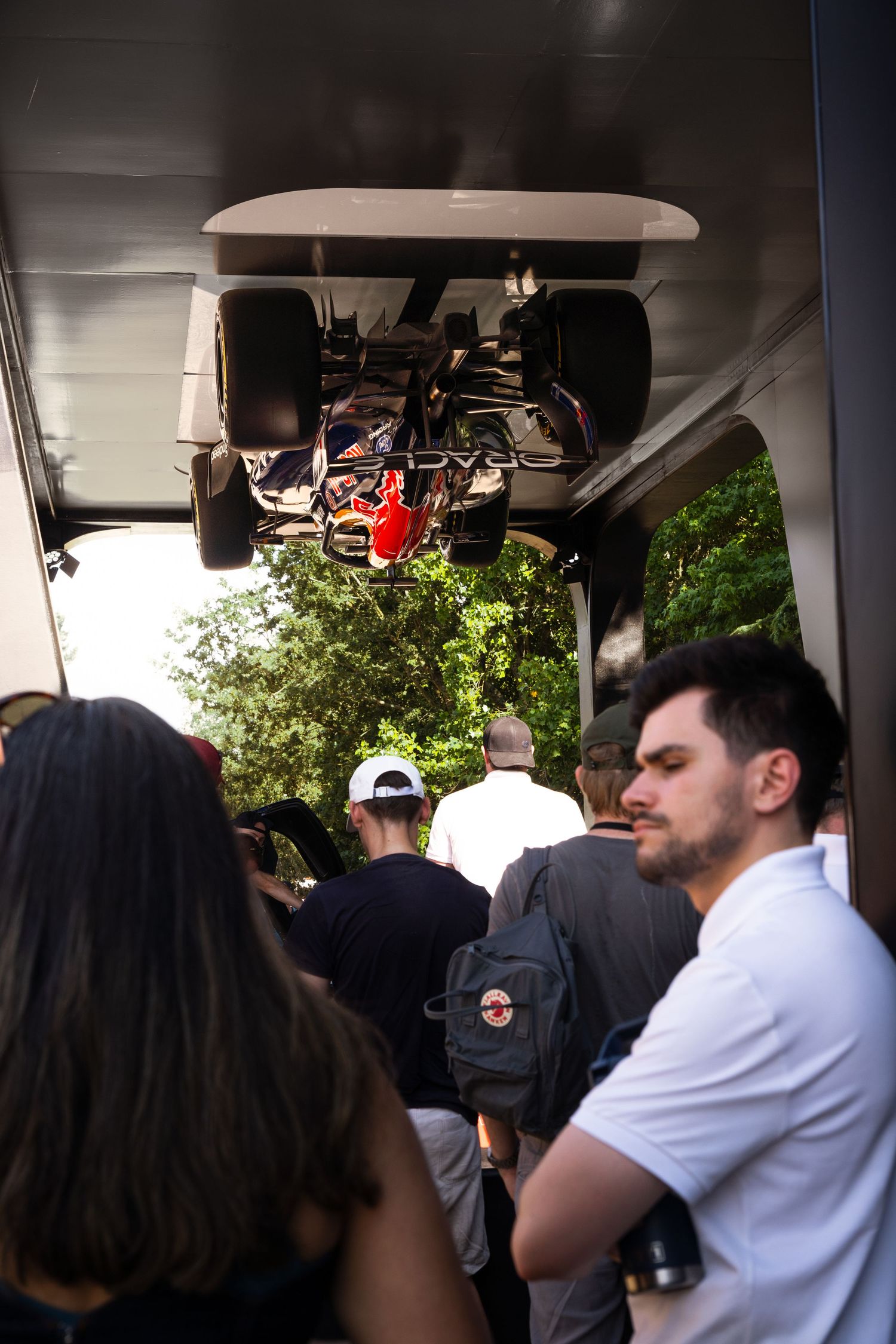 Crowd queuing inside a trailer with an inverted Formula 1 race car displayed on the ceiling at a motorsport event.