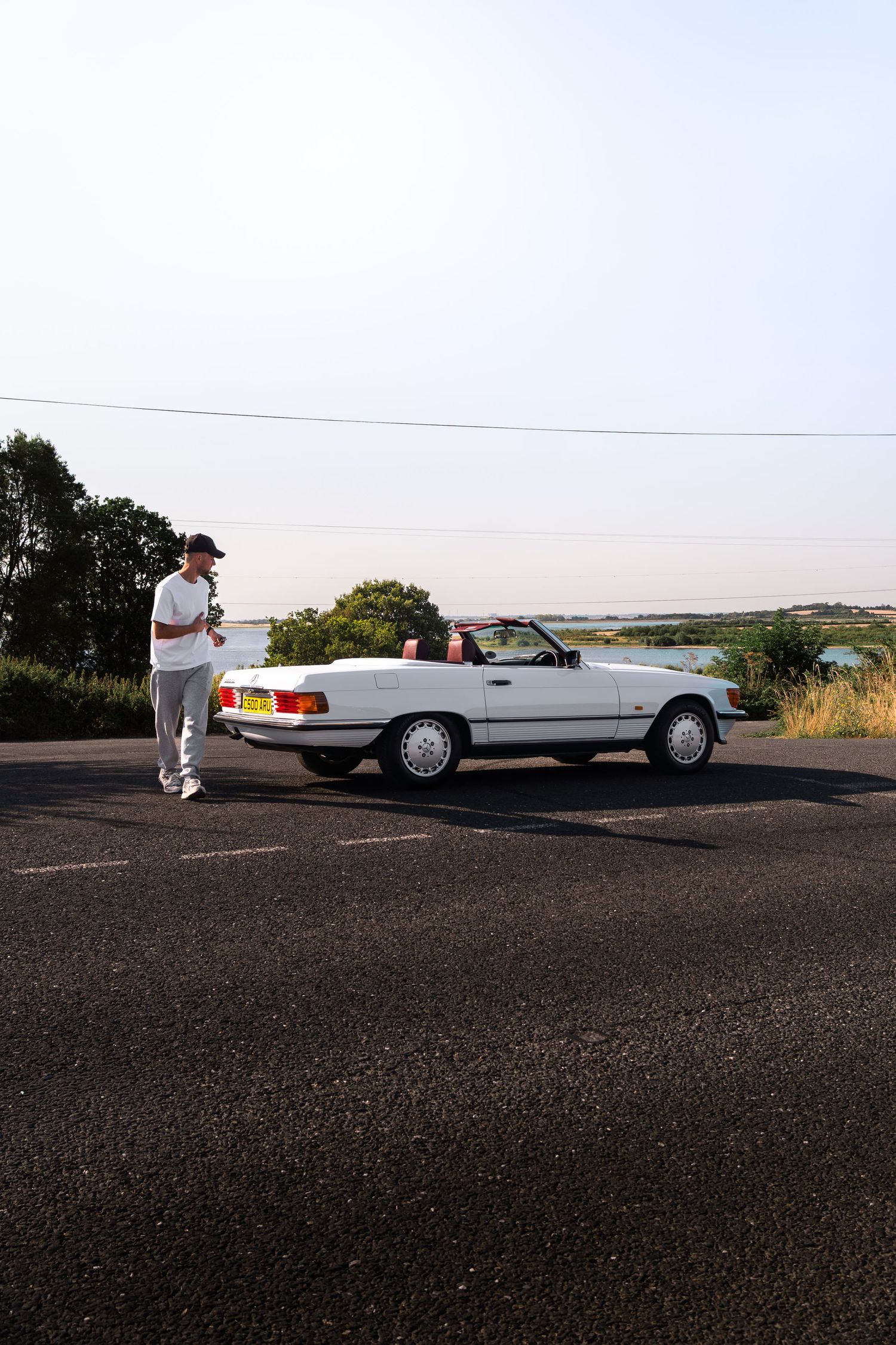 Man standing beside a white Mercedes-Benz convertible parked on a road with scenic water views in the background.