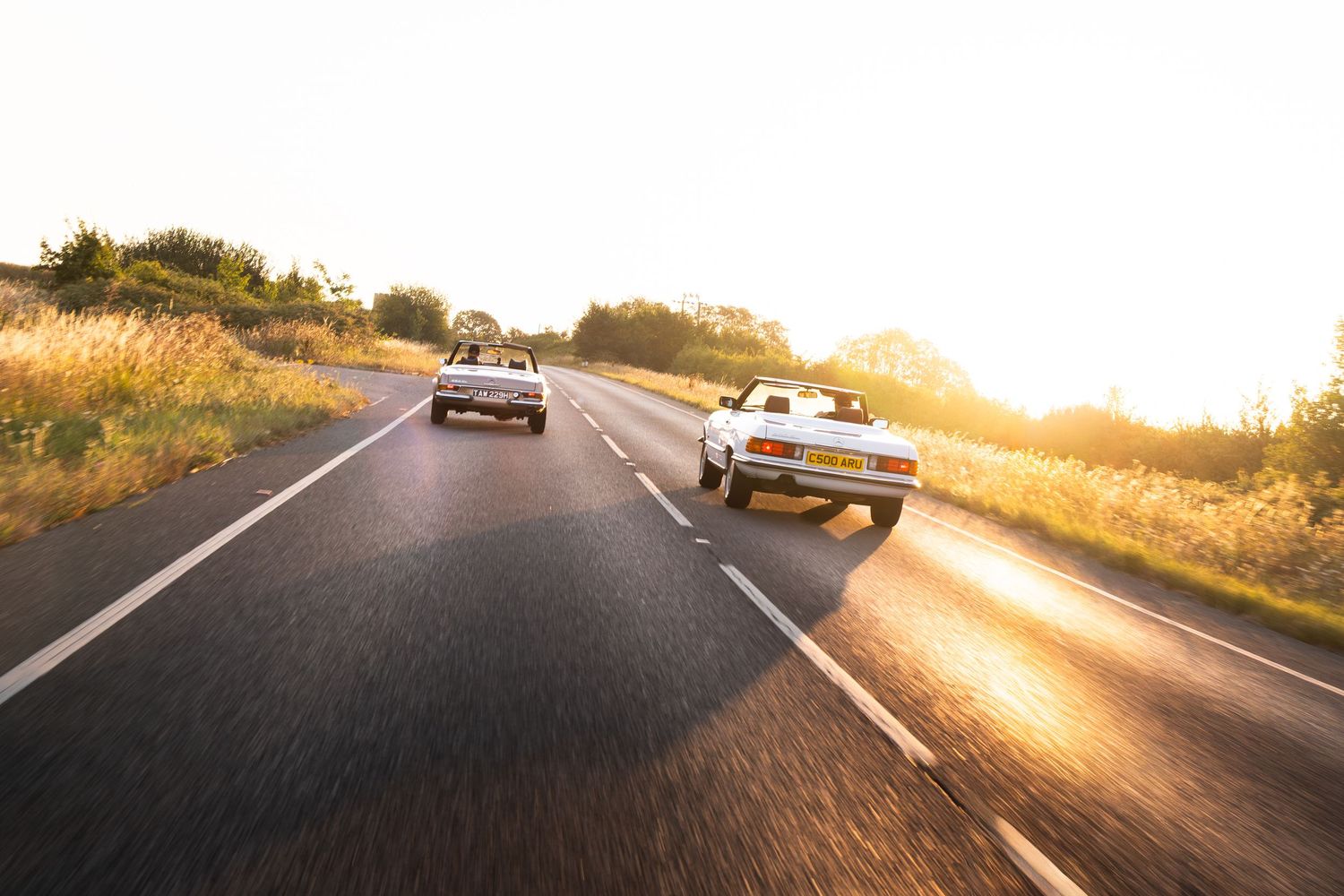 Two classic convertible cars driving on a scenic road at golden sunset hour with warm light and green fields.