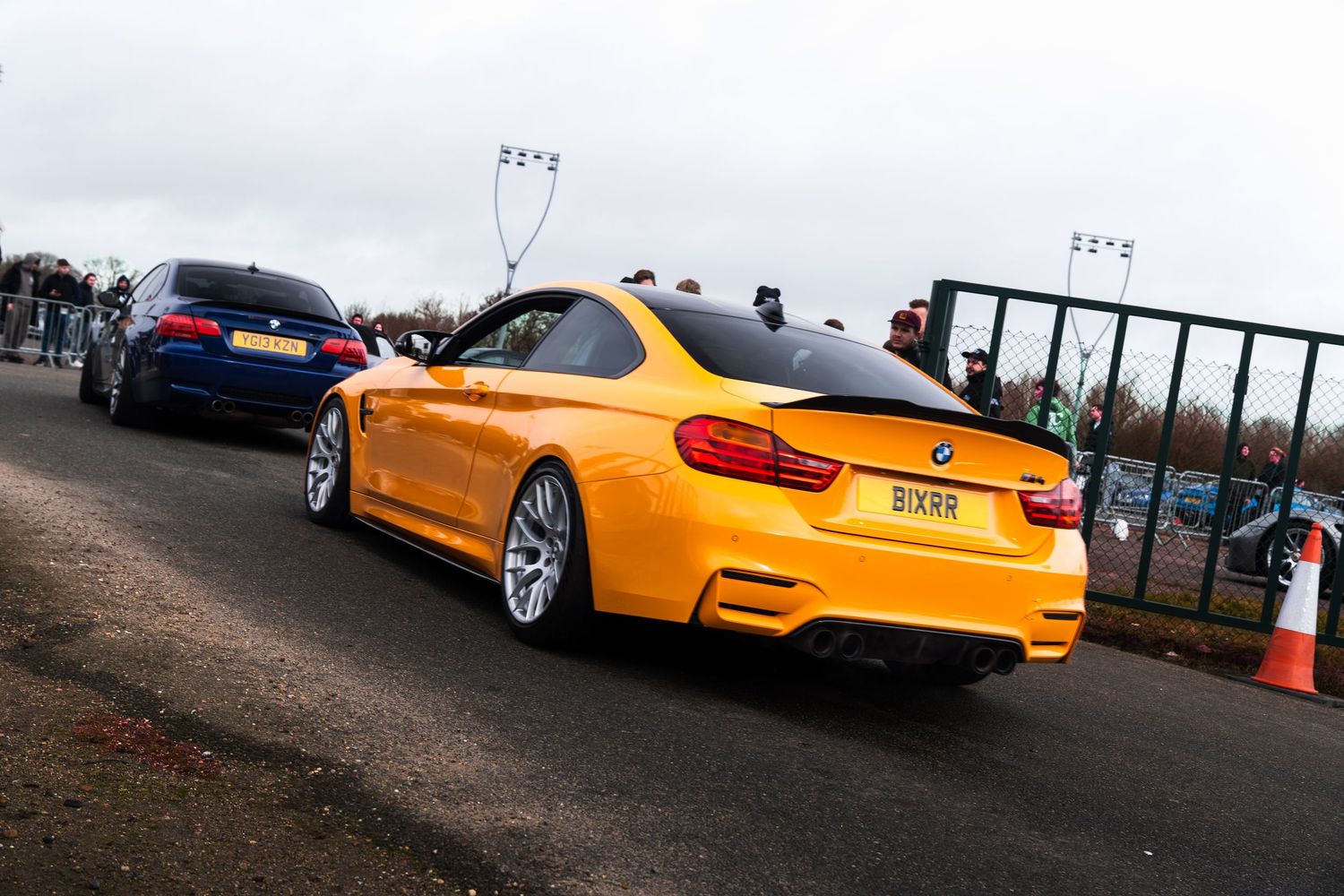 A striking orange BMW M4 coupe parked at an outdoor car meet event with spectators nearby.