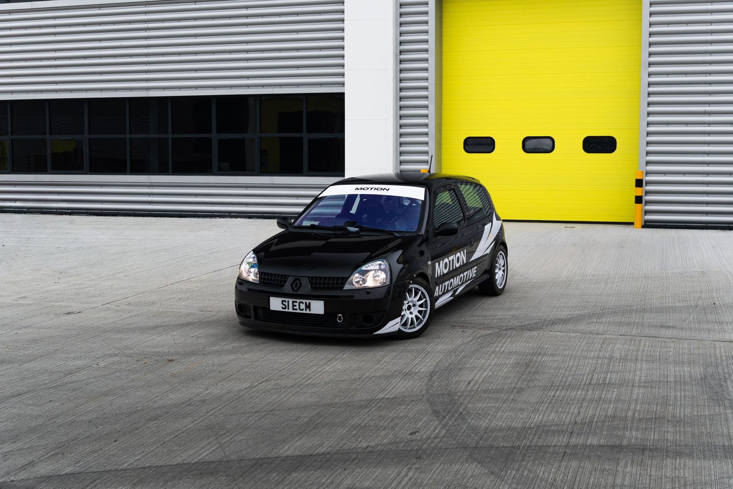 Black Motion Automotive modified Renault Clio hatchback photographed from front angle outside yellow industrial garage.