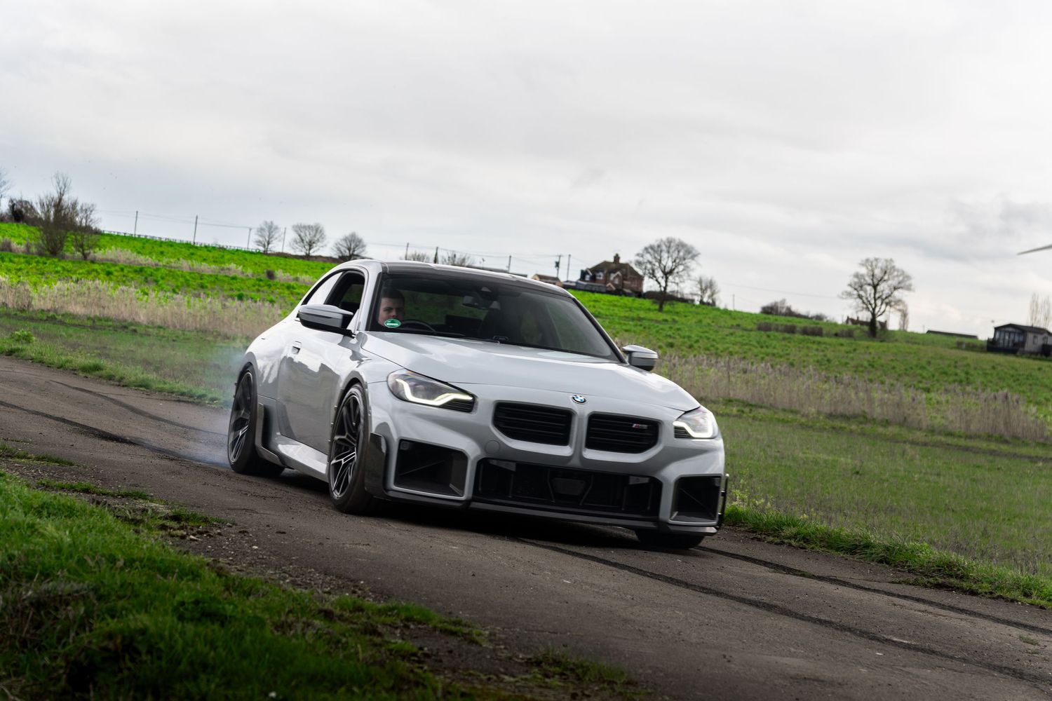 White BMW M2 coupe drifting on a rural road with green hills in the background under cloudy skies.