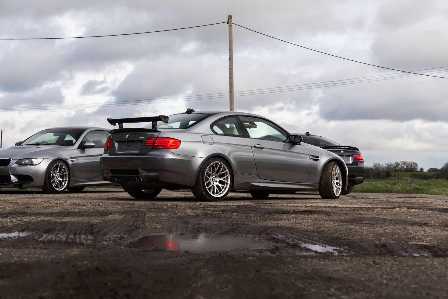 Side rear view of a gray BMW M3 E92 with GT spoiler parked on a muddy gravel lot.