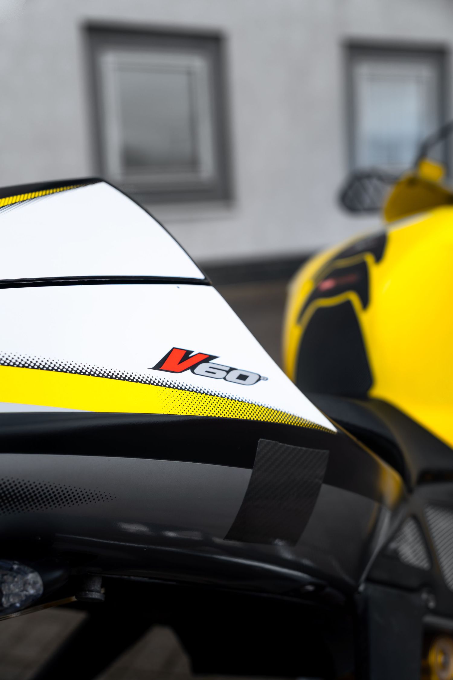 Close-up of Aprilia V60 motorcycle seat fairing showing white and yellow racing livery detail.