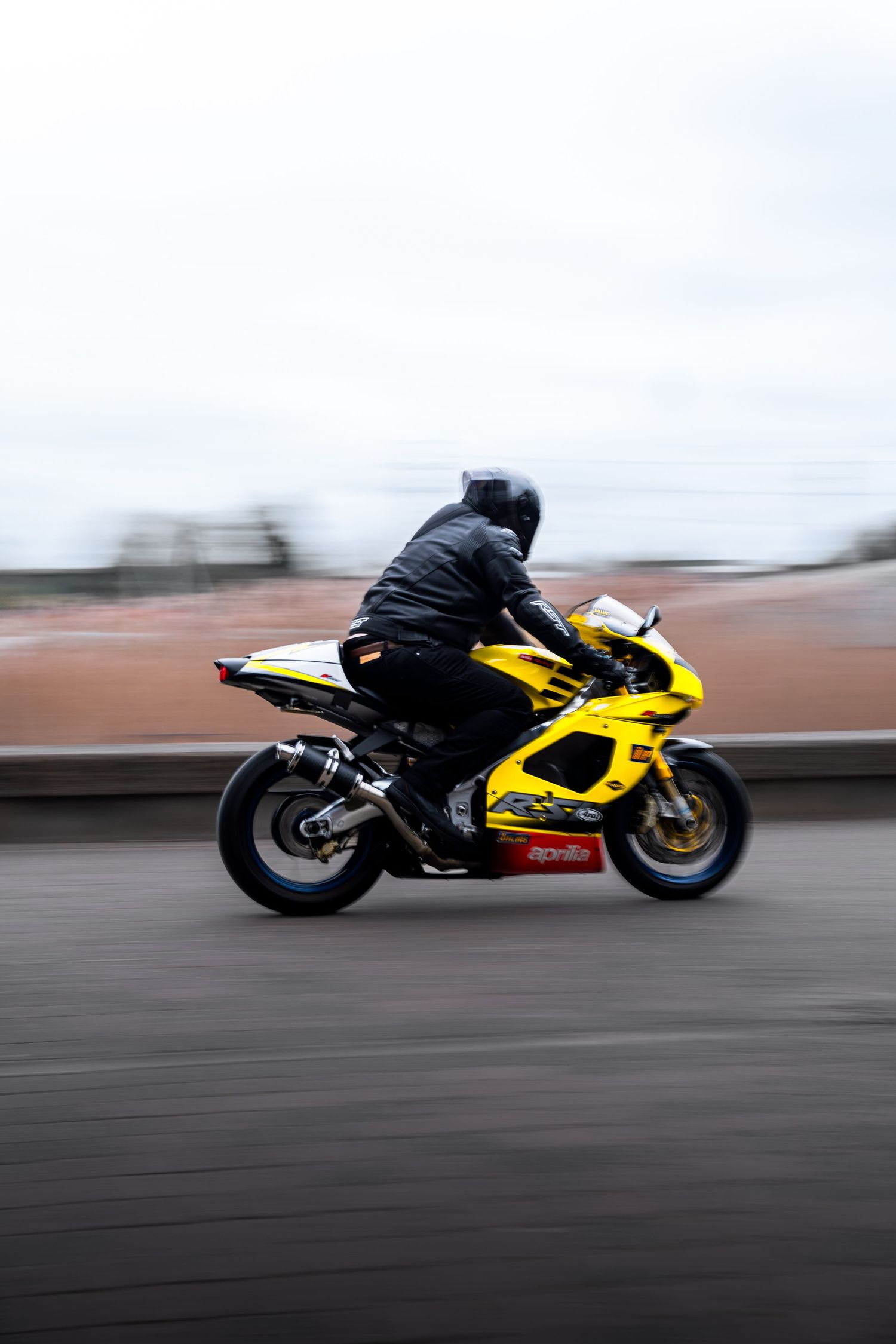 Yellow Aprilia superbike speeding on a road with motion blur effect, ridden by a helmeted rider.