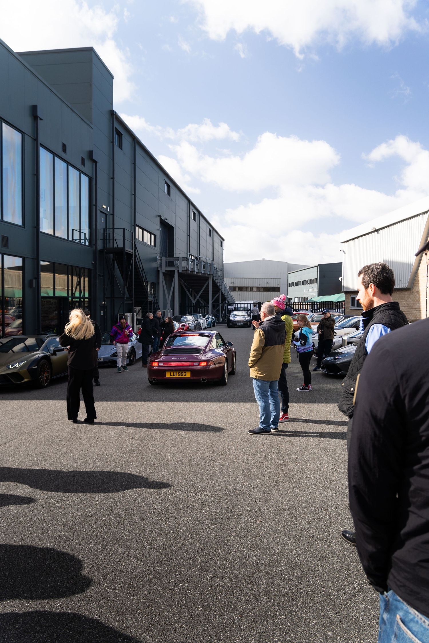 Crowd of car enthusiasts gathered in an industrial parking lot viewing a collection of exotic and sports cars.
