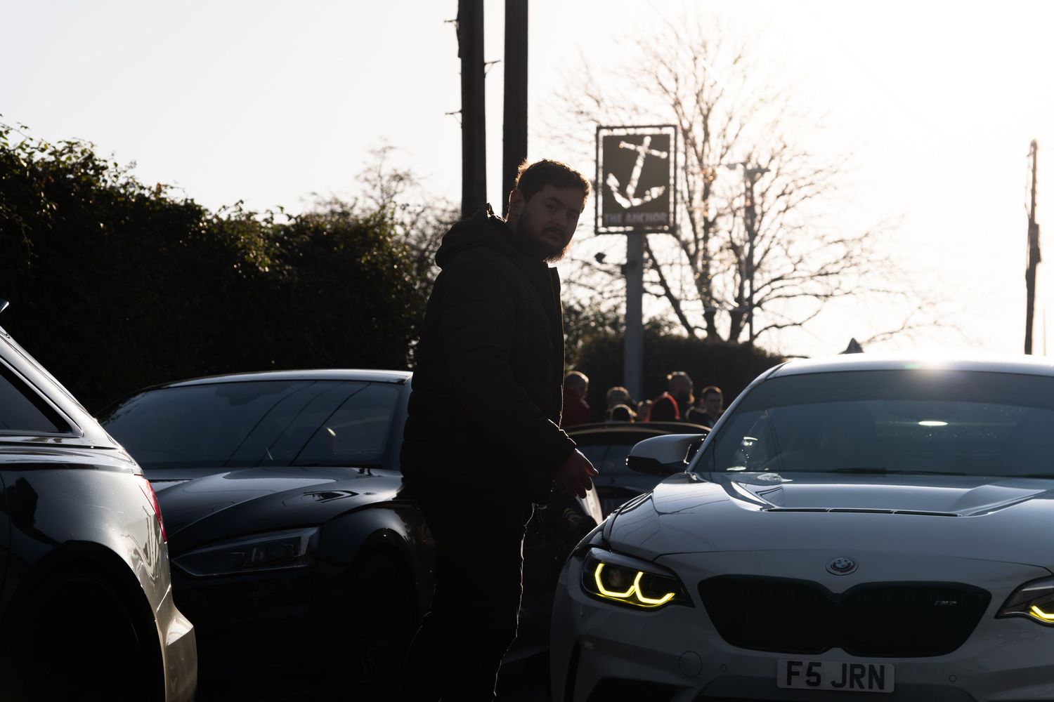 Man standing between cars at a sunny car meet with a BMW M2 featuring angel eyes.