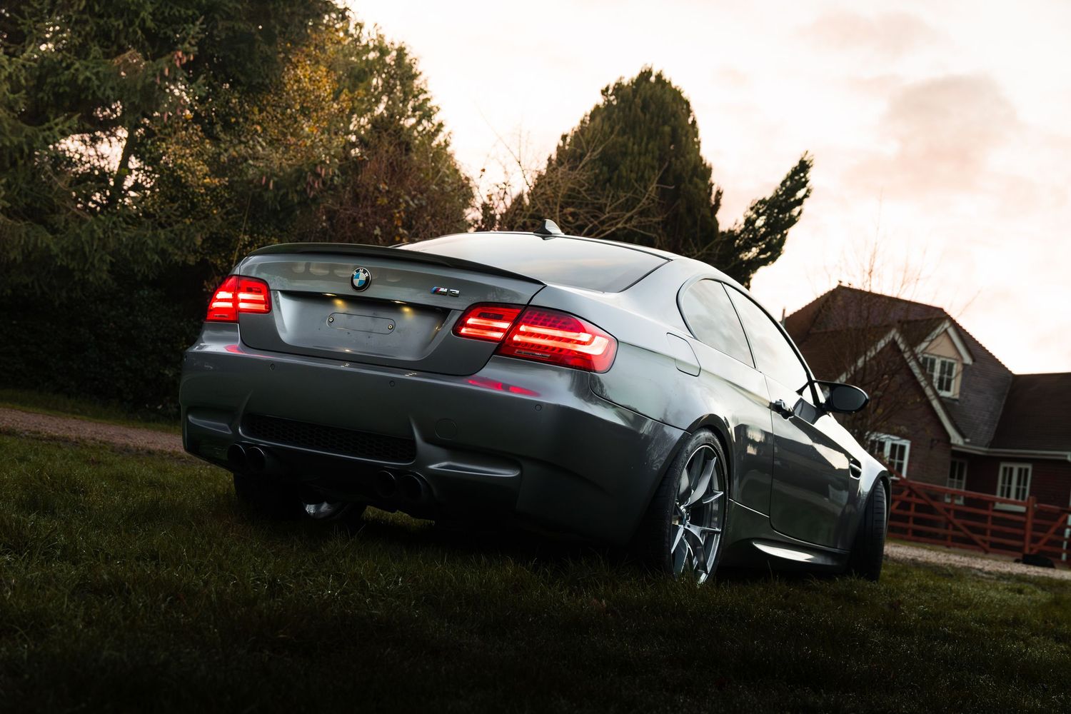 Rear view of grey BMW M3 coupe parked on grass near trees at golden hour.