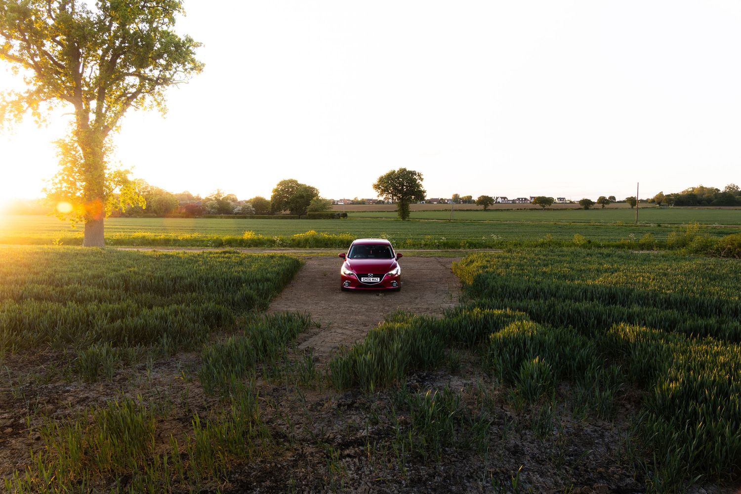 Red car parked on a dirt path in a vast green field during a golden sunset.