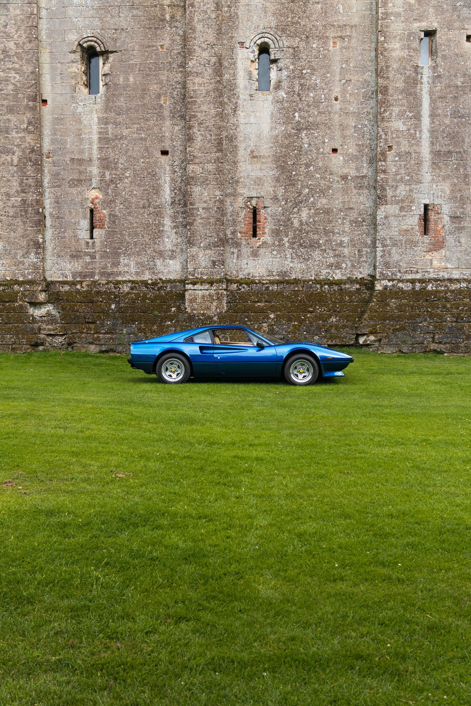 A blue Ferrari 308 GTS parked on green grass in front of an ancient stone castle wall.