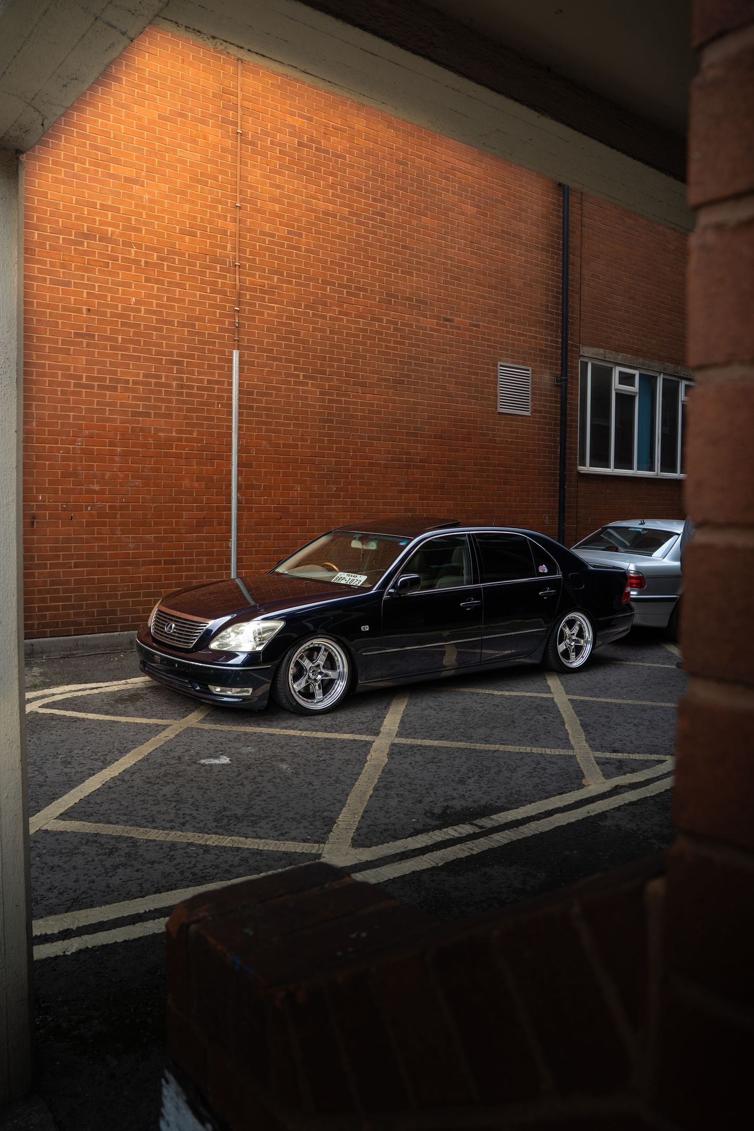 Black Lexus LS sedan parked in a brick-walled car park with yellow box junction markings.