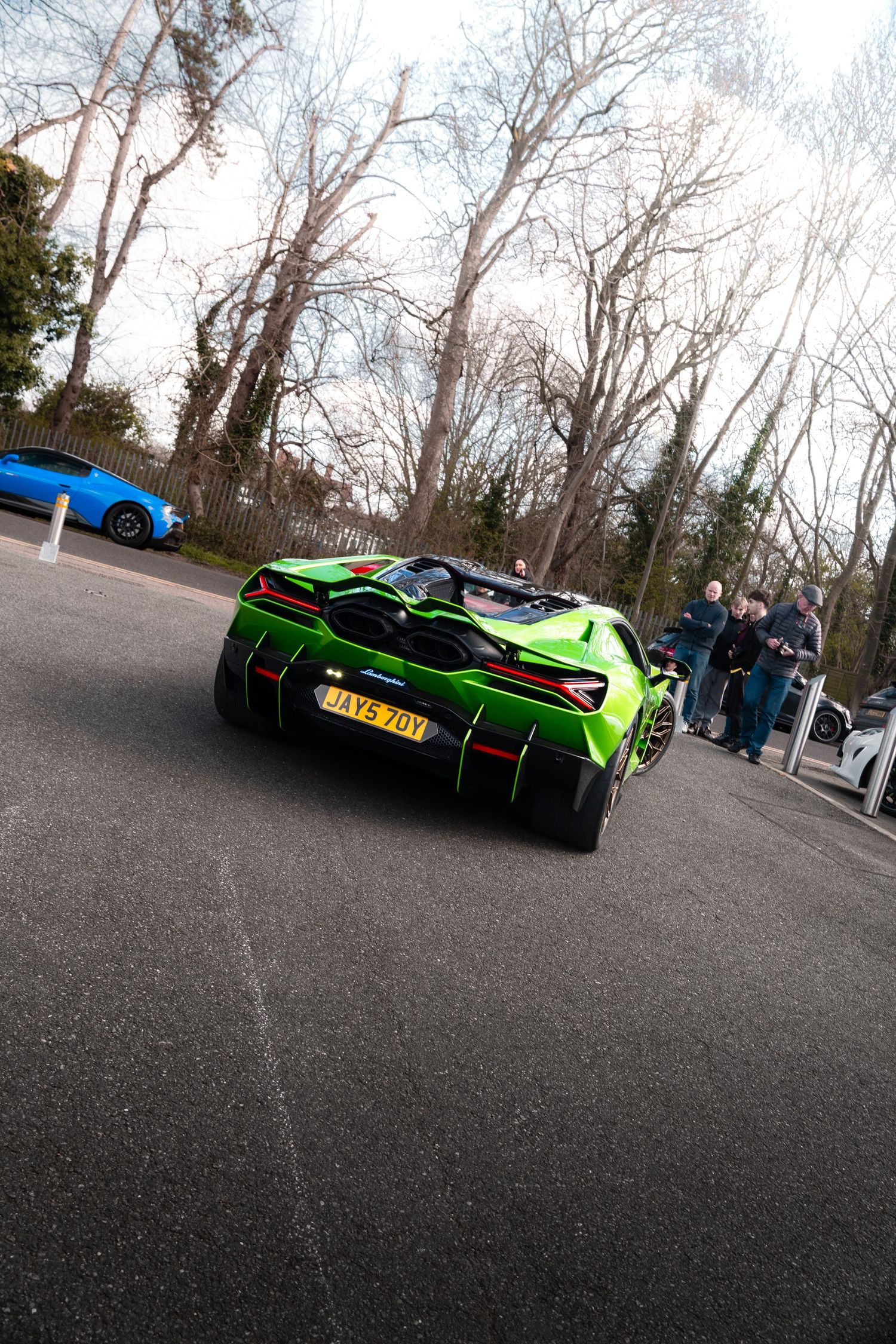 Green Lamborghini Sian rear view on a road with spectators and bare trees in background.