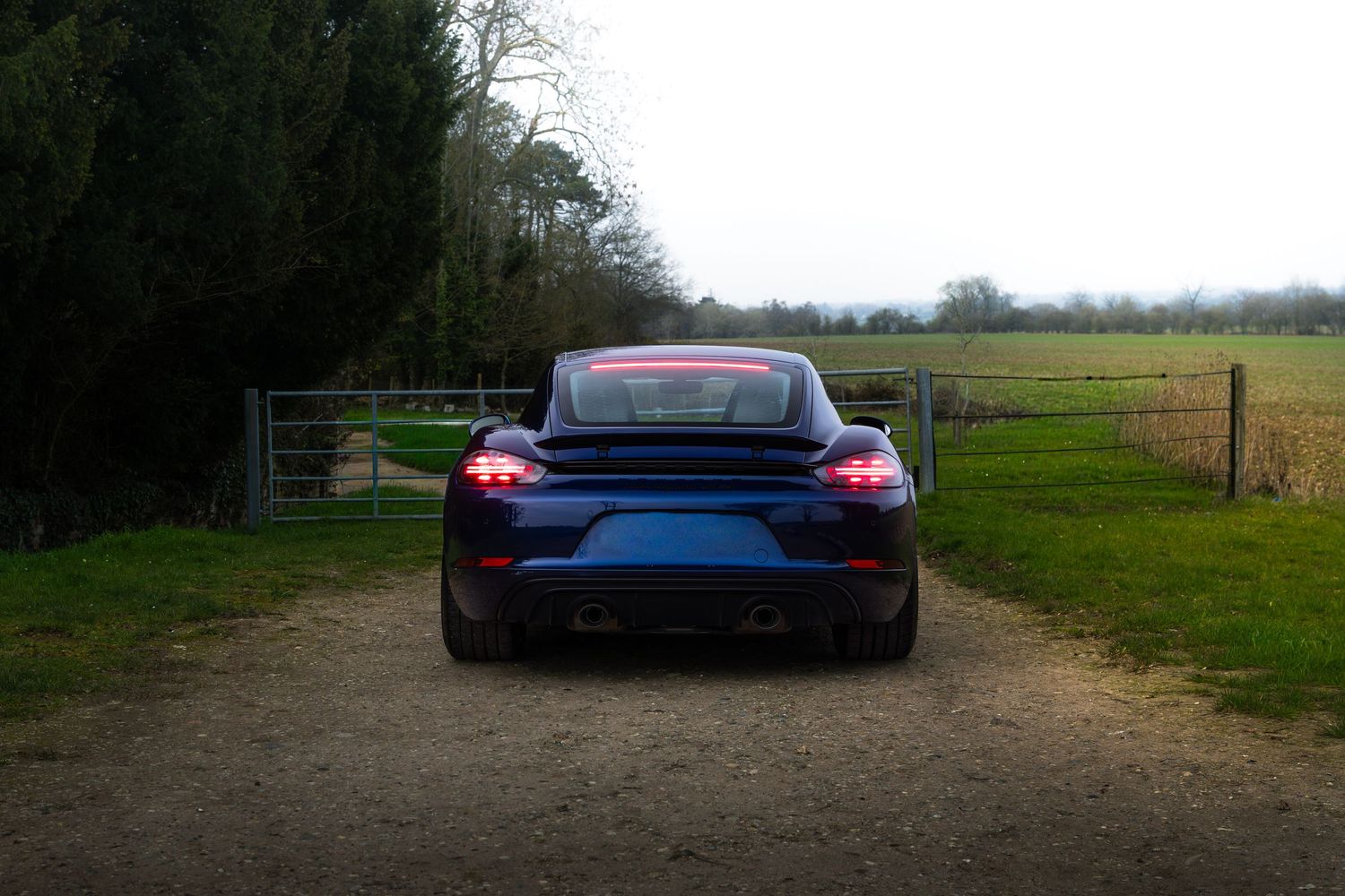 Rear of a blue Porsche 718 Cayman with red brake lights glowing near a countryside farm gate.