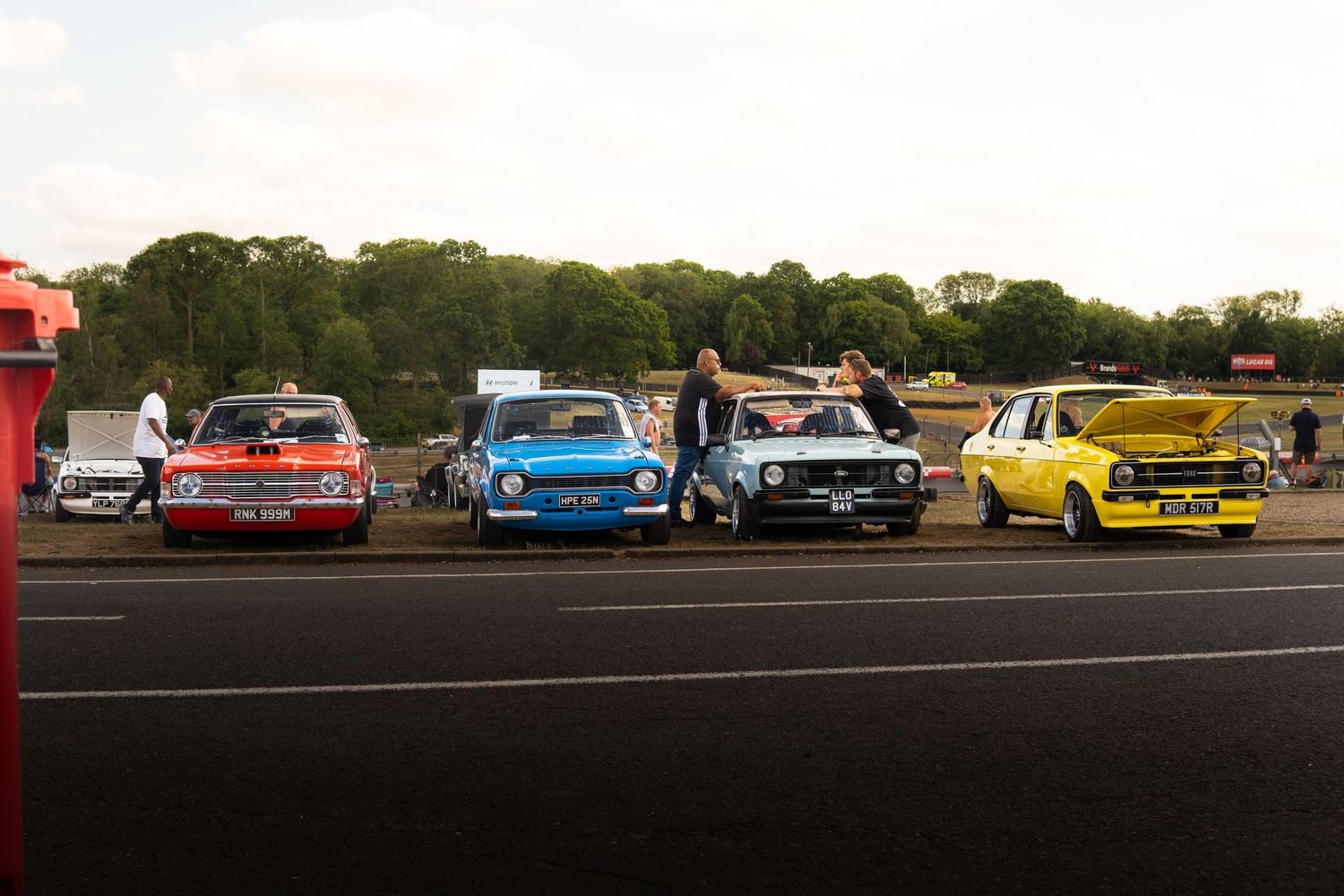 Vintage classic cars lined up on a racetrack at dusk with spectators nearby.