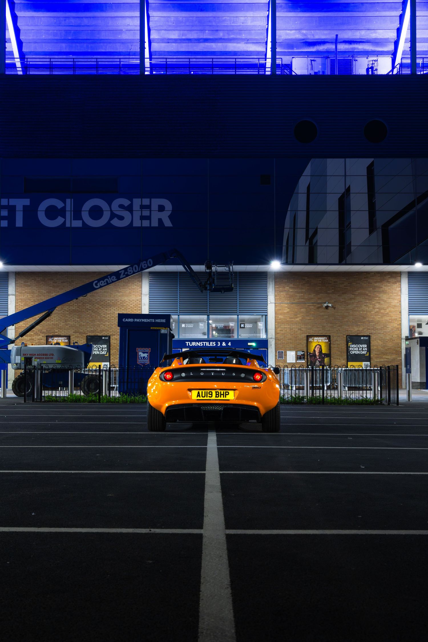 Rear view of orange Lotus Elise AU19BHP parked facing blue-lit stadium turnstiles entrance at night.