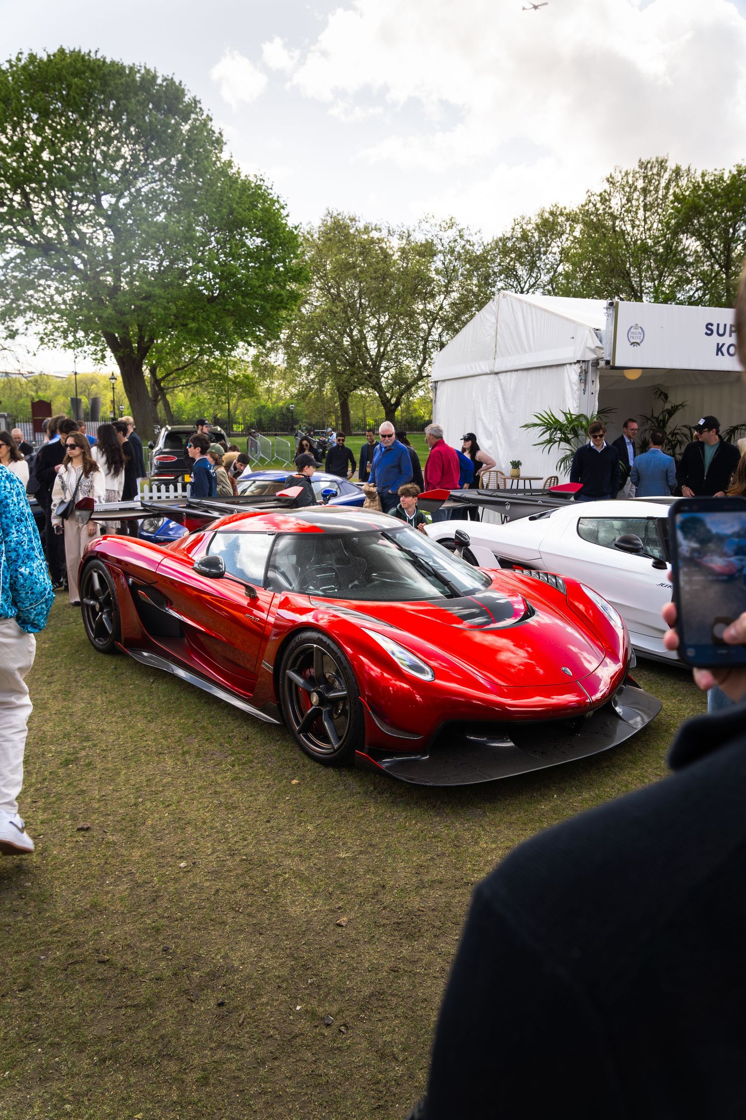 A stunning red Koenigsegg Agera hypercar on display at an outdoor luxury supercar show with crowds.
