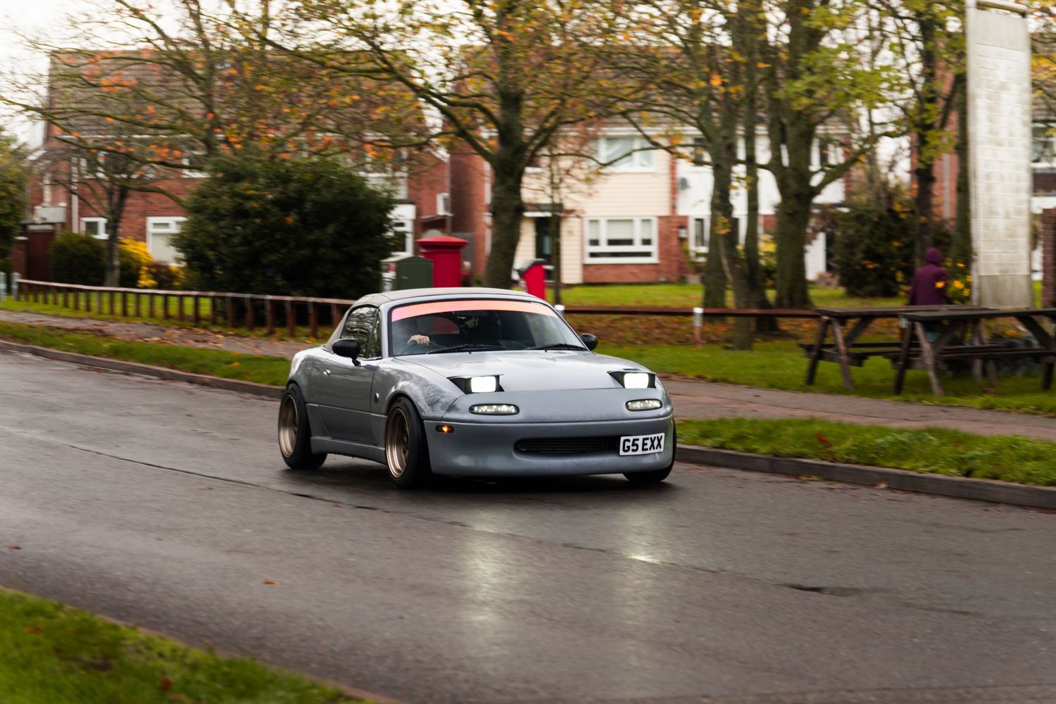 Modified grey Mazda MX-5 Miata driving on wet residential street.