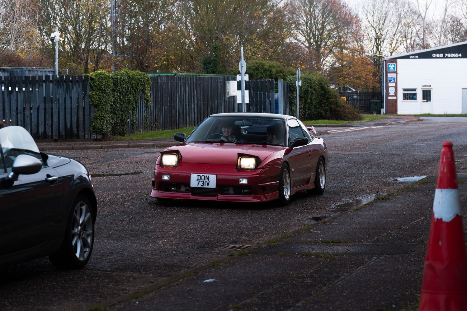 A red classic sports car with headlights on drives through a car park surrounded by cones.